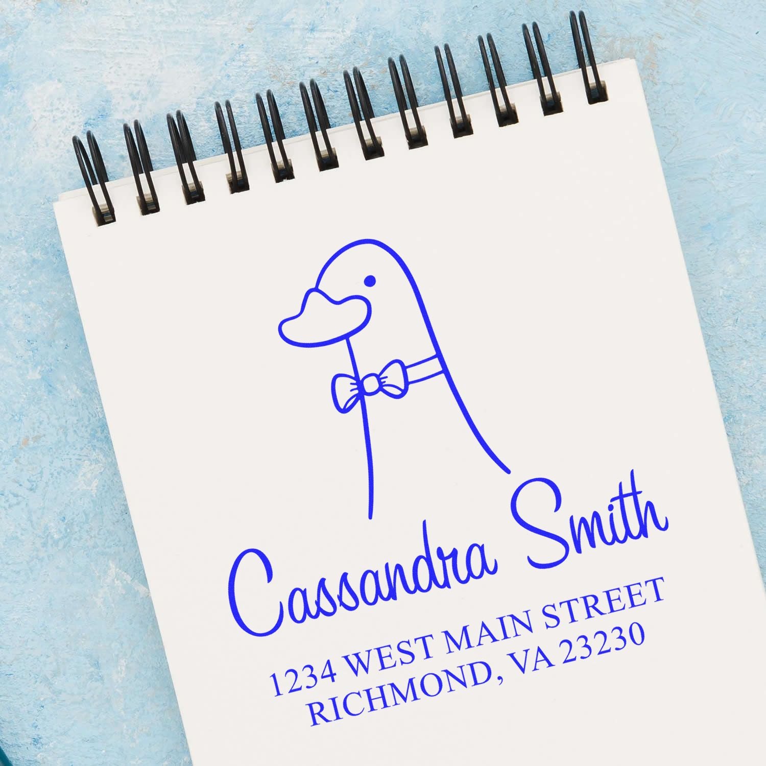 A notebook displays a stamped image of a duck with a bow tie, accompanied by the name "Cassandra Smith" and an address. The Portable Pre-Inked Dashing Duck Made To Order Address Stamp is used as a Custom Home Stamp on the page.