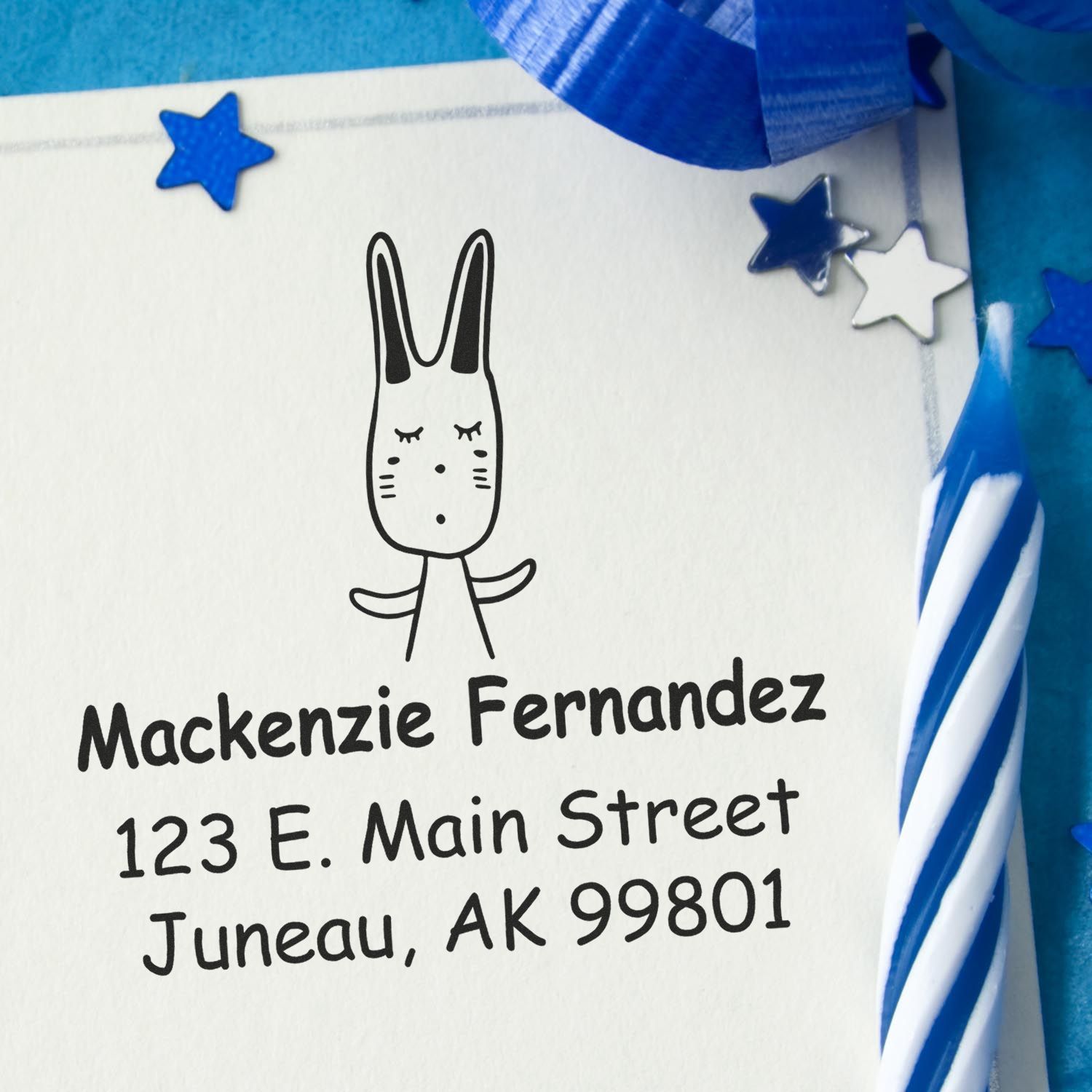 Wood Handle Hopping Bunny Cartoon Animal Custom Return Address Stamper on an envelope with "Mackenzie Fernandez, 123 E. Main Street, Juneau, AK 99801." Personalizable New Address Stamp beside a blue and white candle and star decorations.