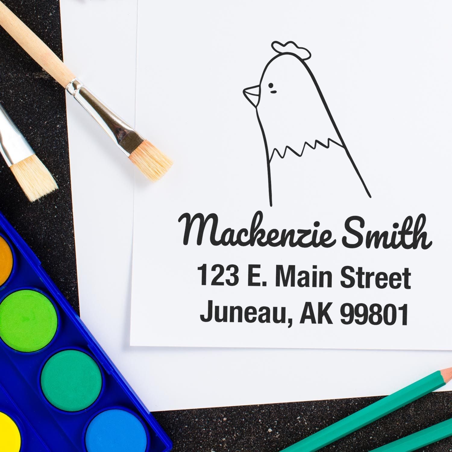Wood handle clucking chicken cartoon animal custom home address stamp on paper with personalized address stamp reading "Mackenzie Smith, 123 E. Main Street, Juneau, AK 99801." Surrounded by paintbrushes and watercolor paints.