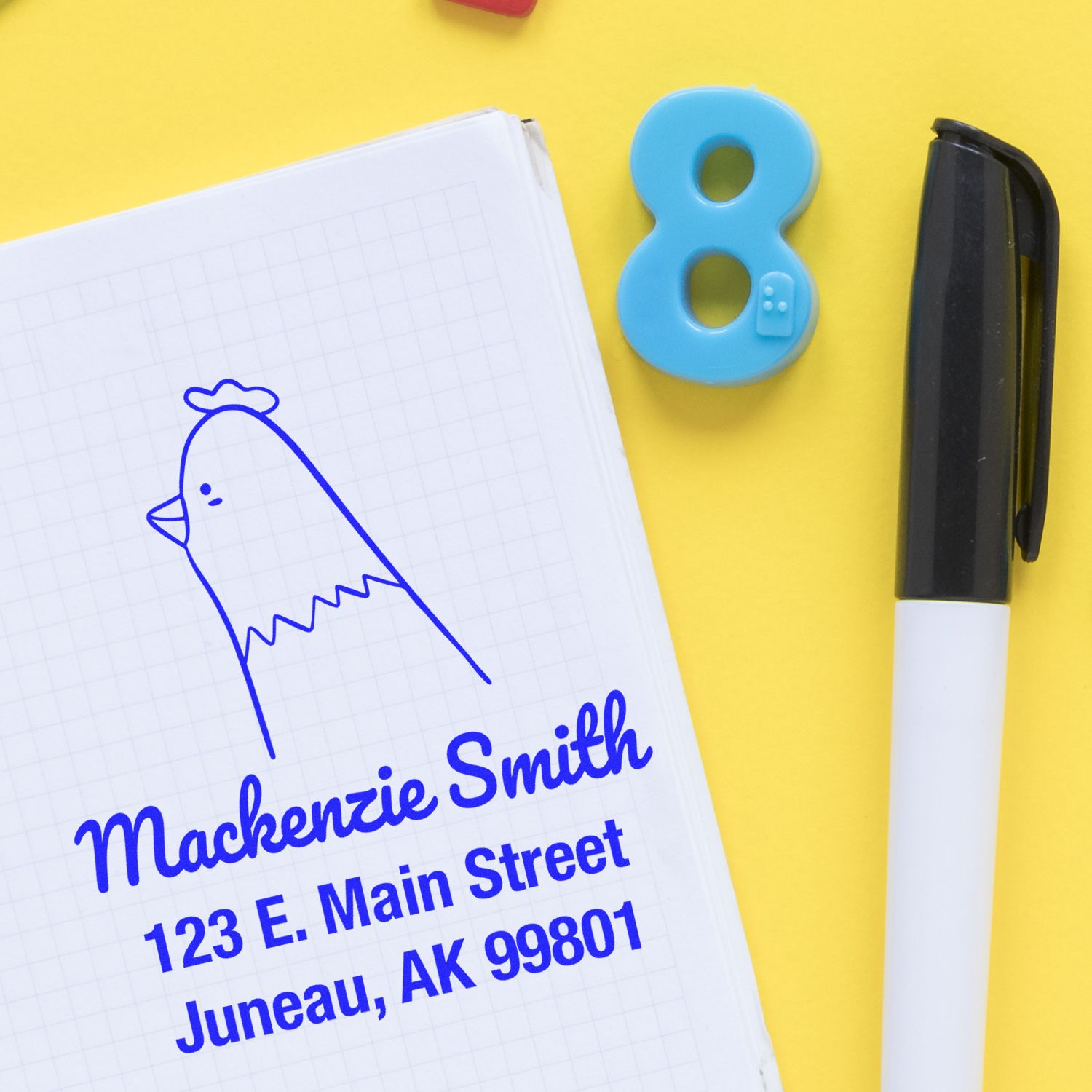 Image of a Portable Pre-Inked Clucking Chicken Made To Order Name And Address Stamper on a notepad. The customizable address stamp displays "Mackenzie Smith, 123 E. Main Street, Juneau, AK 99801" in blue ink, with a pen and number eight nearby on a yellow background.
