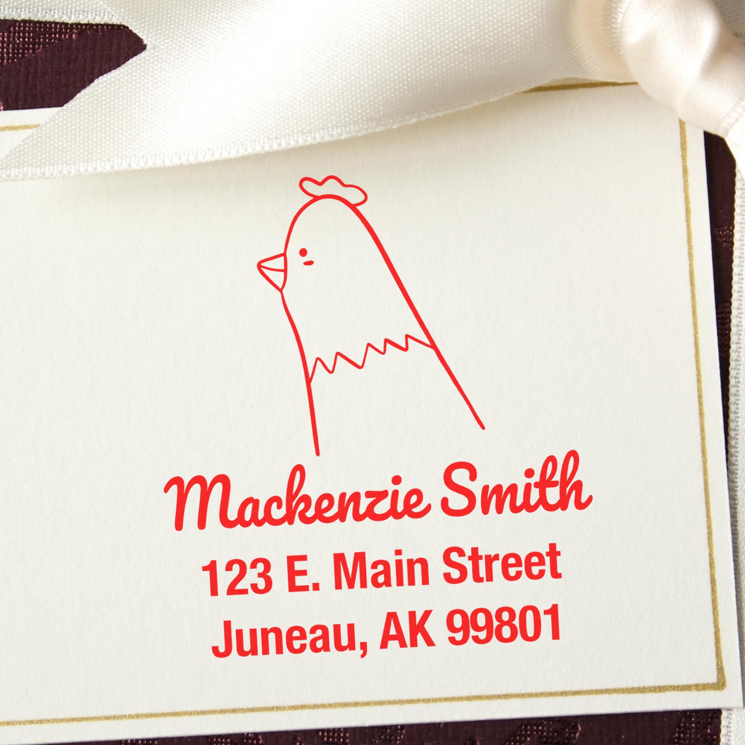 Portable Pre-Inked Clucking Chicken Made To Order Name And Address Stamper in use, featuring a red chicken design with the text "Mackenzie Smith, 123 E. Main Street, Juneau, AK 99801." Ideal for a Customized Mailing Stamp on a cream card with a white ribbon.