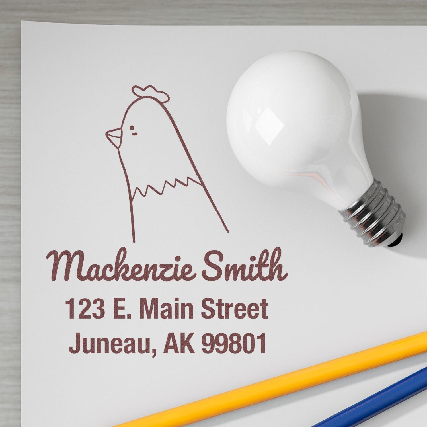 Wood handle clucking chicken cartoon animal custom home address stamp on paper with "Mackenzie Smith, 123 E. Main Street, Juneau, AK 99801." Personalized new address stamp next to a light bulb and pencils on a desk.