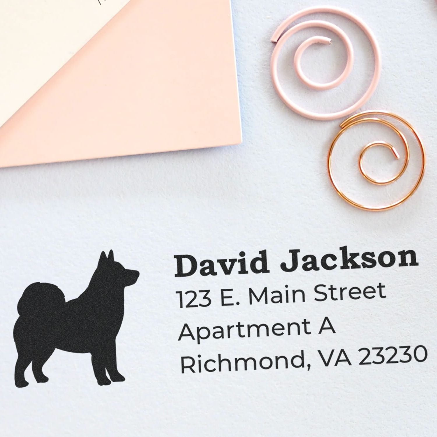 Self-Inking Alaskan Malamute Dog Address Stamp on a letter, featuring a silhouette of the dog and sample address text. Nearby are pink and gold spiral paper clips on a light background.