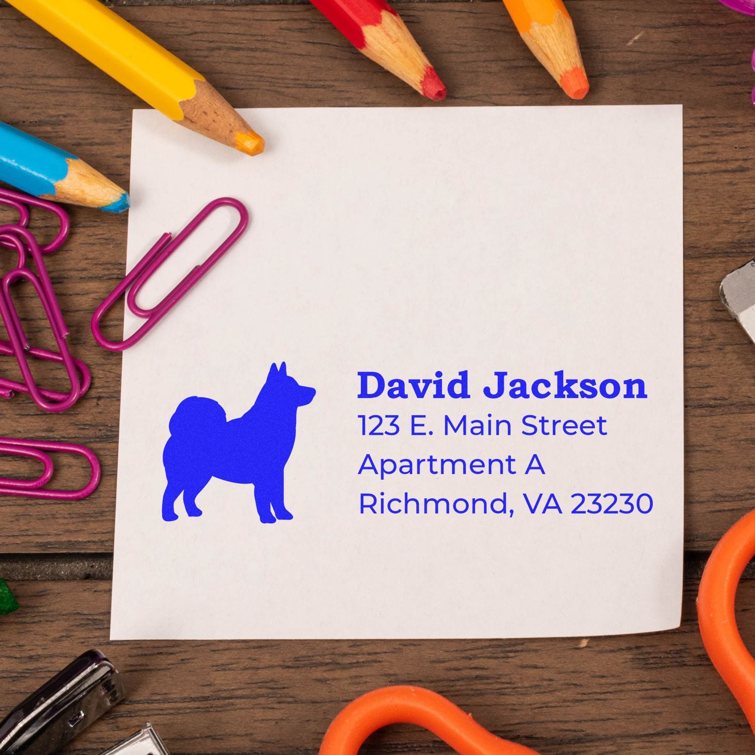 PSI Pre-Inked Personalized Alaskan Malamute Address Stamp on paper, surrounded by colorful pencils and paperclips on a wooden desk.