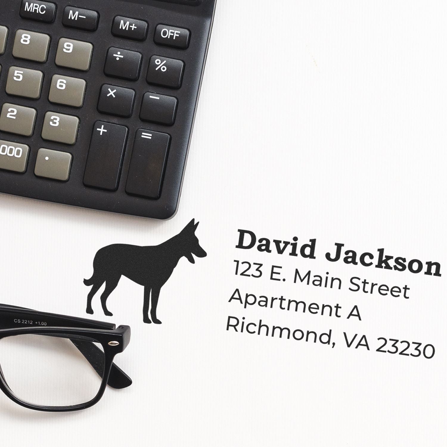 PSI Pre-Inked Personalized Belgian Malinois Address Stamp on paper next to a calculator and glasses, featuring a silhouette of a Belgian Malinois and sample address text.