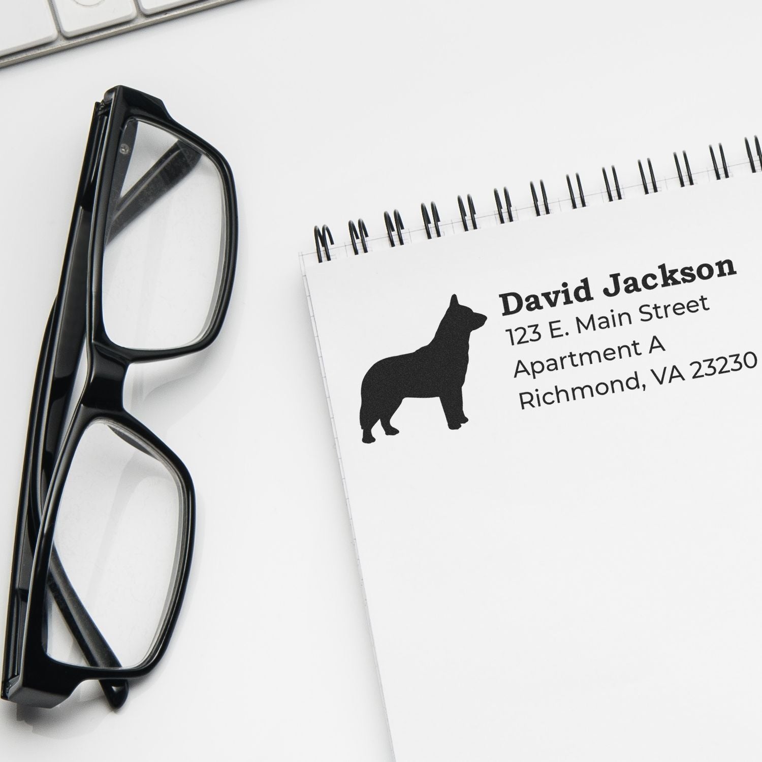 A notebook with an imprint from the Self-Inking Blue Heeler Dog Address Stamp, featuring a silhouette of a dog next to an address. Glasses and a keyboard are nearby.