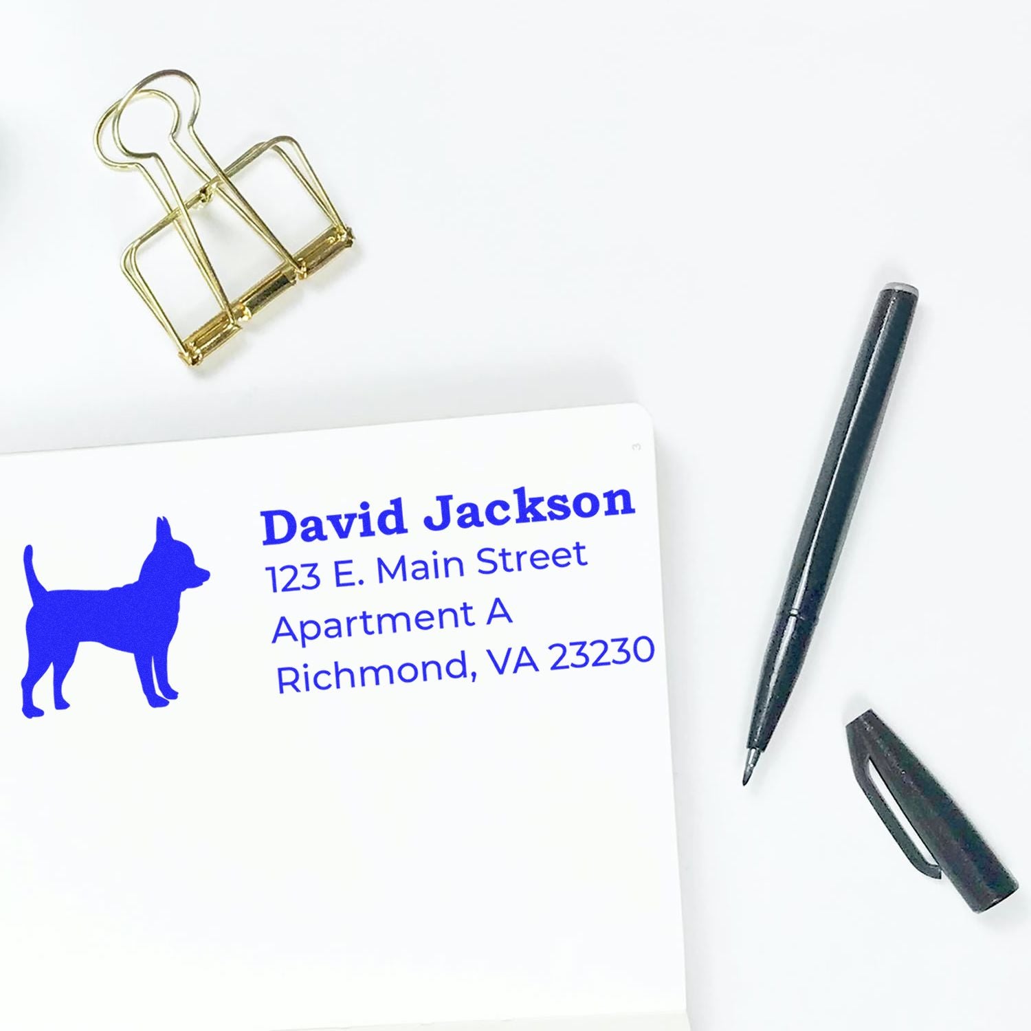 Self-Inking Chihuahua Dog Address Stamp on white paper with blue text, featuring a Chihuahua silhouette. Nearby are a black pen, cap, and a gold binder clip.