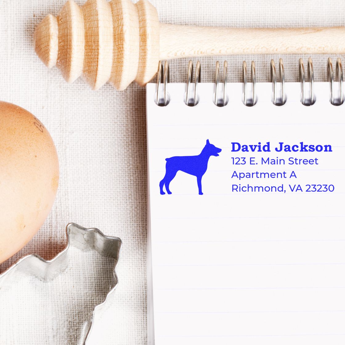 A notepad displays a blue Doberman Silhouette Address Rubber Stamp with sample text. Nearby are a honey dipper, egg, and cookie cutter on a textured surface.