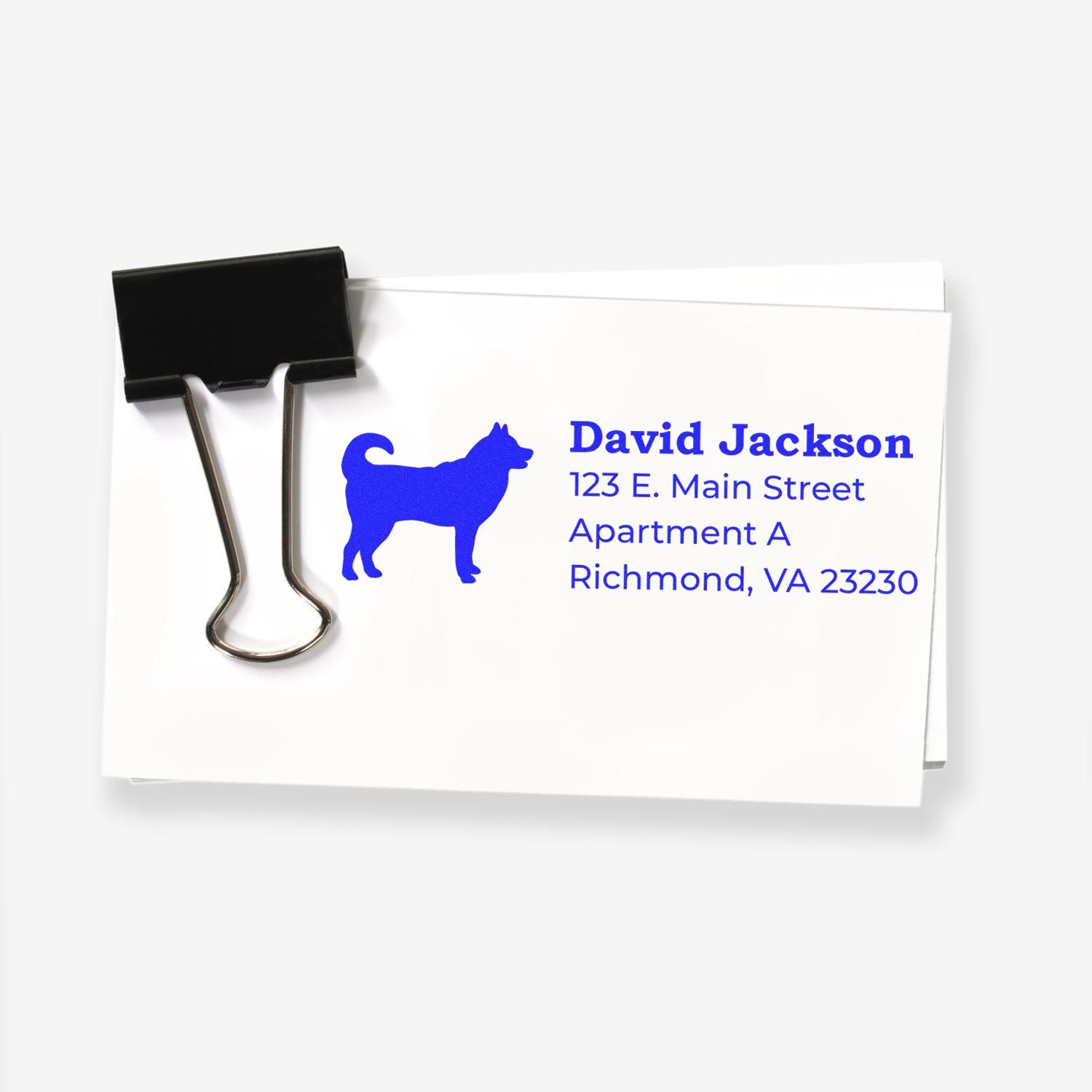 Slim Pre-Inked Husky Address Stamp on white paper with a blue husky silhouette and sample address. A black binder clip holds the paper stack.