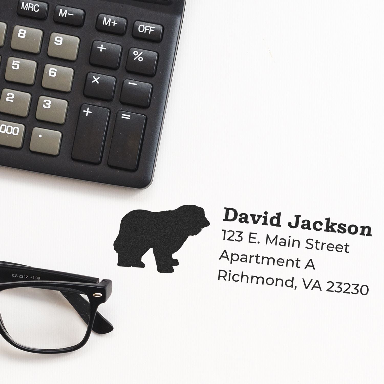 A Slim Pre-Inked Old English Sheepdog Address Stamp on white paper next to a calculator and glasses, featuring a silhouette of a sheepdog and printed address details.