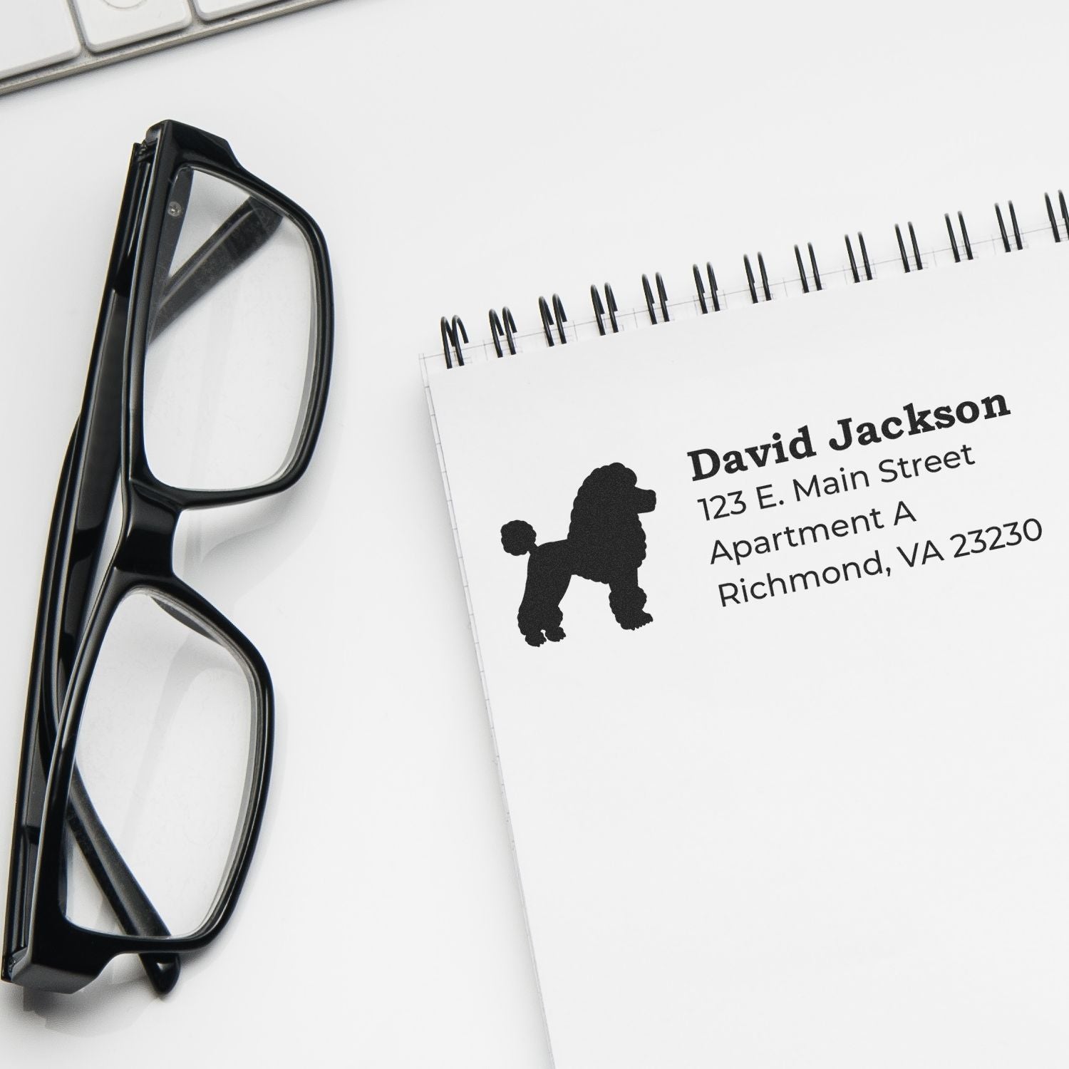 A notepad with a Poodle Silhouette Address Rubber Stamp imprint, showing a poodle silhouette next to an address. Glasses and a keyboard are nearby, emphasizing a professional desk setup.