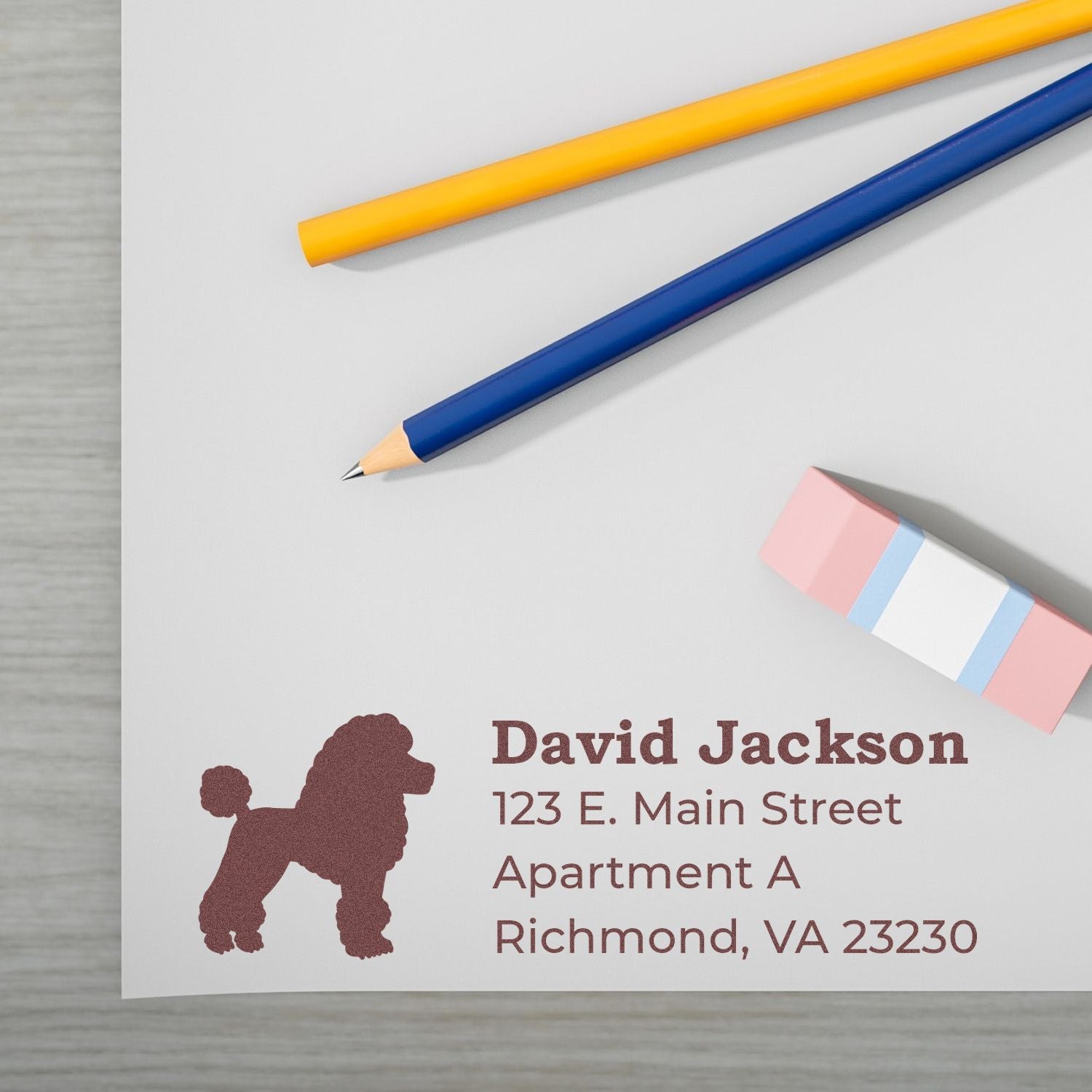 Self-Inking Poodle Dog Address Stamp on white paper with a brown poodle silhouette, next to colorful pencils and an eraser. Text reads David Jackson, 123 E. Main Street, Richmond, VA 23230.