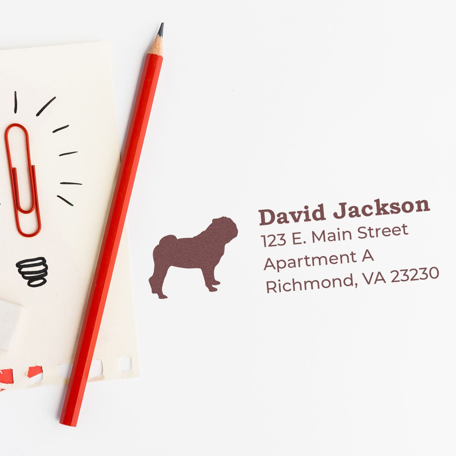 Slim Pre-Inked Pug Address Stamp on paper with a red pencil and paperclip. The stamp features a pug silhouette and sample address text in brown ink.