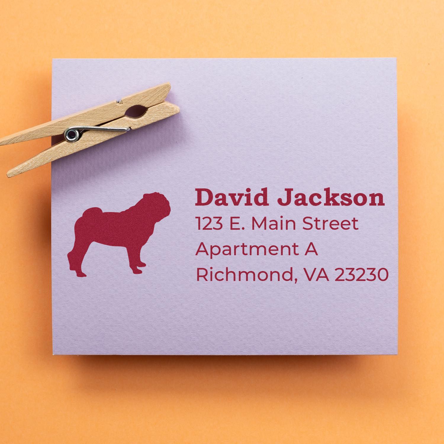 PSI Pre-Inked Personalized Pug Address Stamp on a lavender envelope with a red pug silhouette and address details, clipped with a wooden clothespin on an orange background.