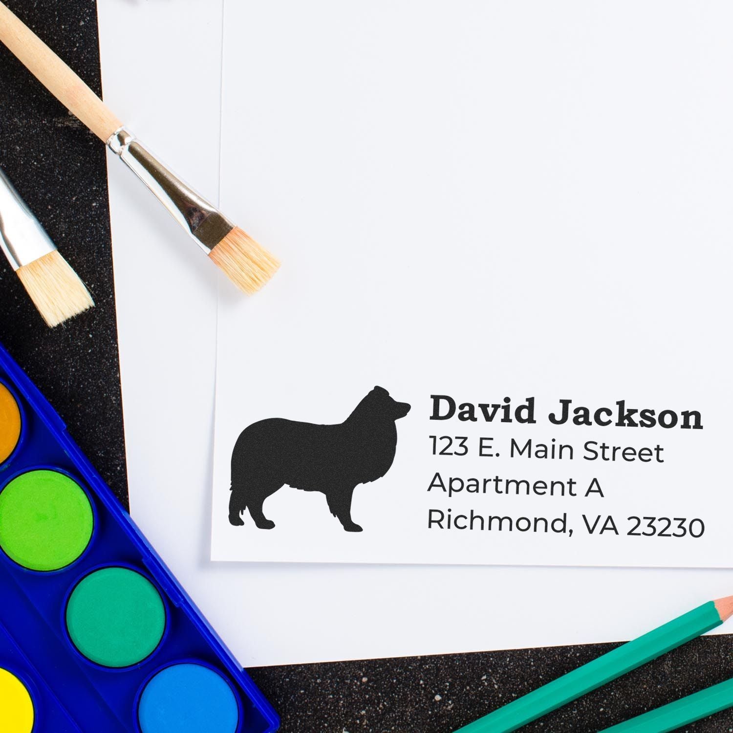 PSI Pre-Inked Personalized Shetland Sheepdog Address Stamp on white paper, featuring a silhouette of the dog. Nearby are paintbrushes and watercolor paints on a black surface.