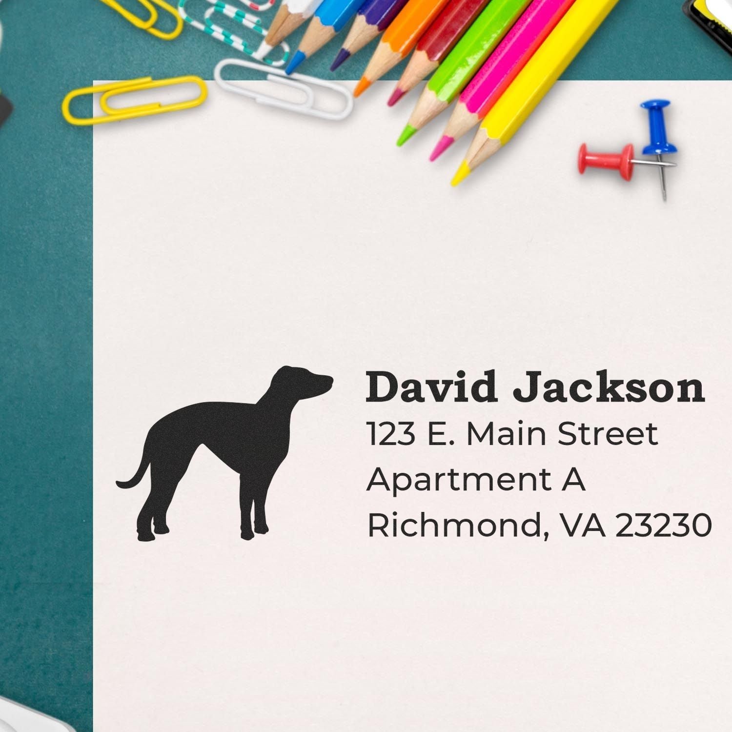 Slim Pre-Inked Whippet Address Stamp on paper with a silhouette of a whippet, surrounded by colorful pencils, paperclips, and push pins on a green surface.