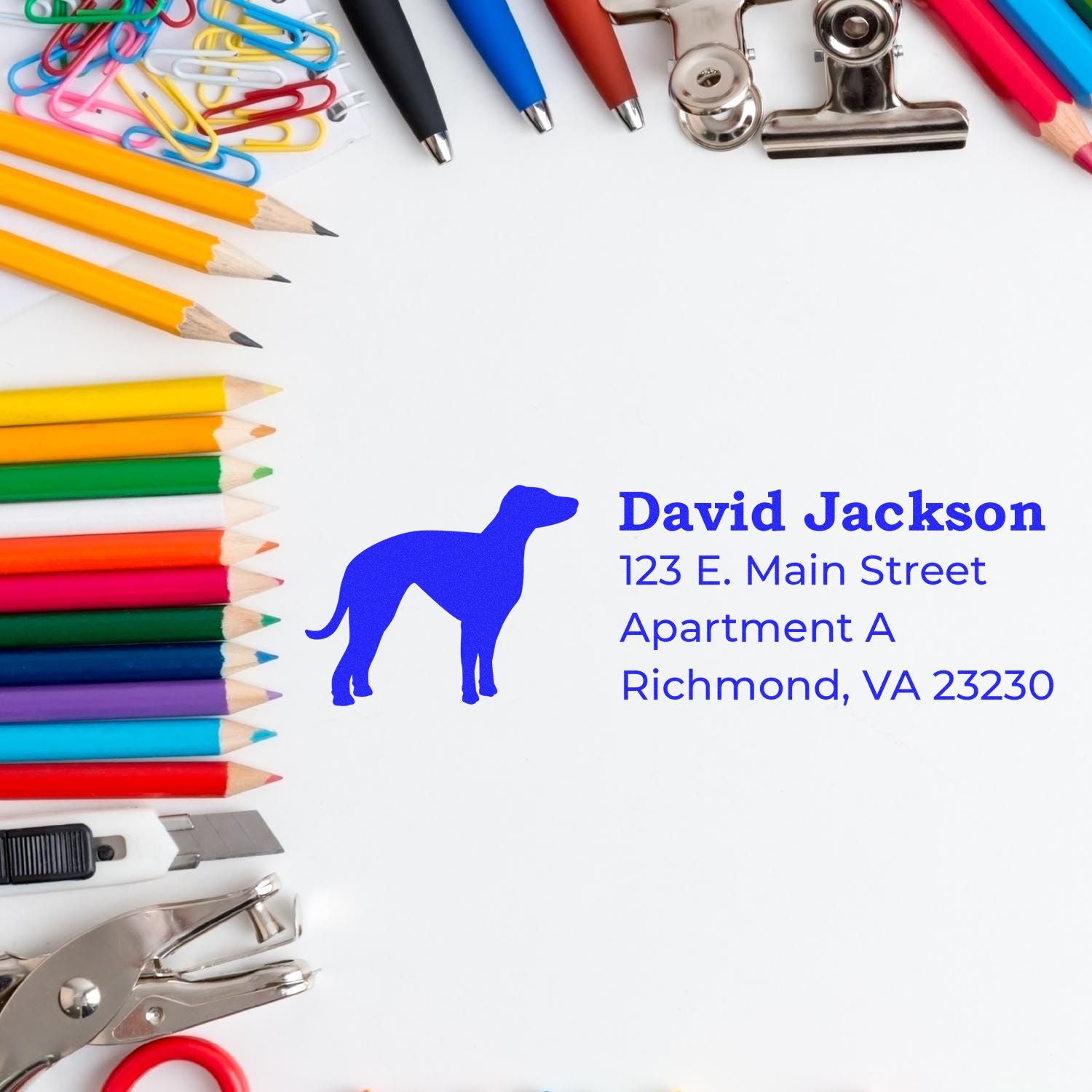 A Whippet Silhouette Address Rubber Stamp on white paper, surrounded by colorful pencils, paperclips, and office supplies. The stamp displays a whippet silhouette with sample address text.