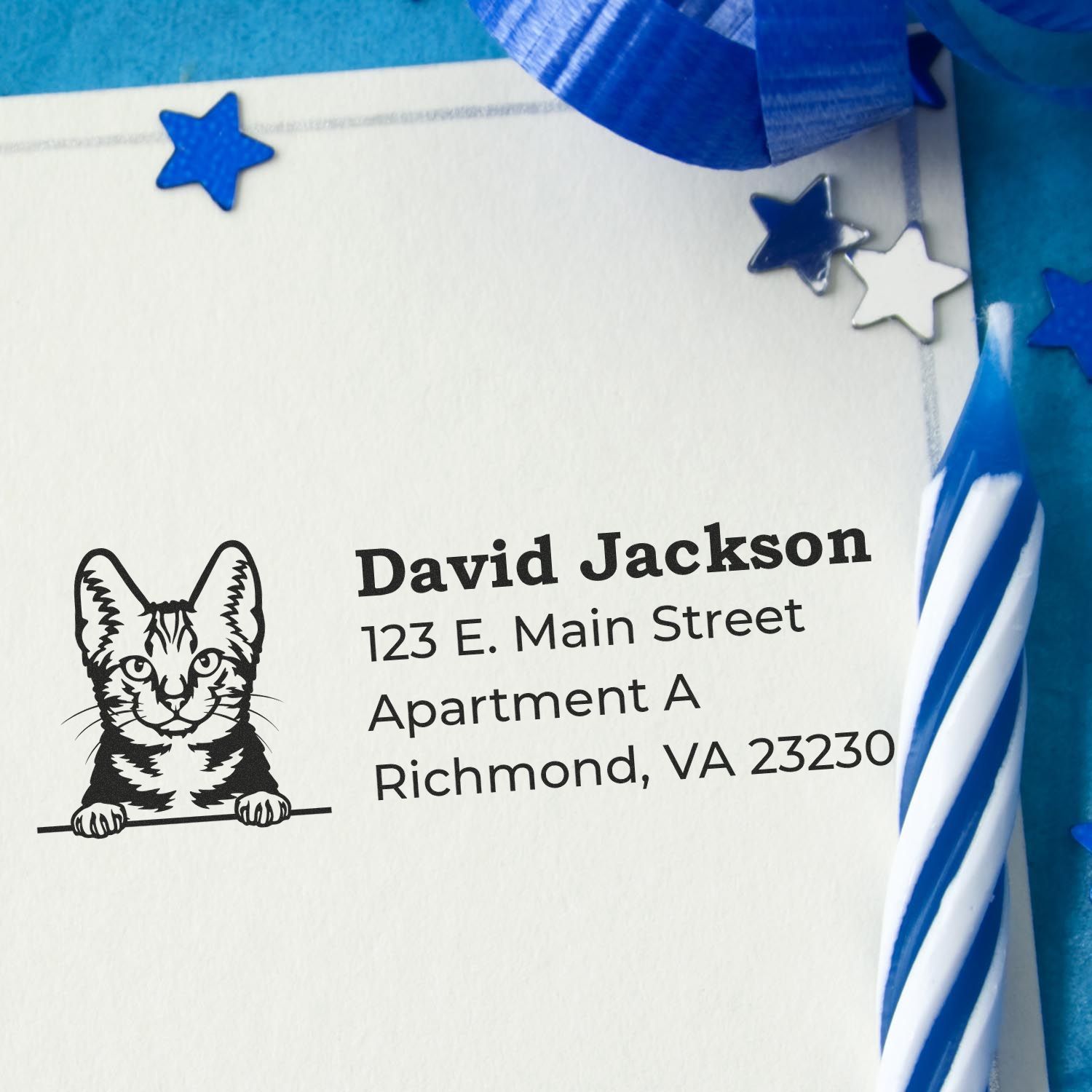 Slim Pre-Inked American Shorthair Peeking Cat Return Address Stamp on an envelope with blue decorations and a striped candle, showcasing a playful cat design above the address text.