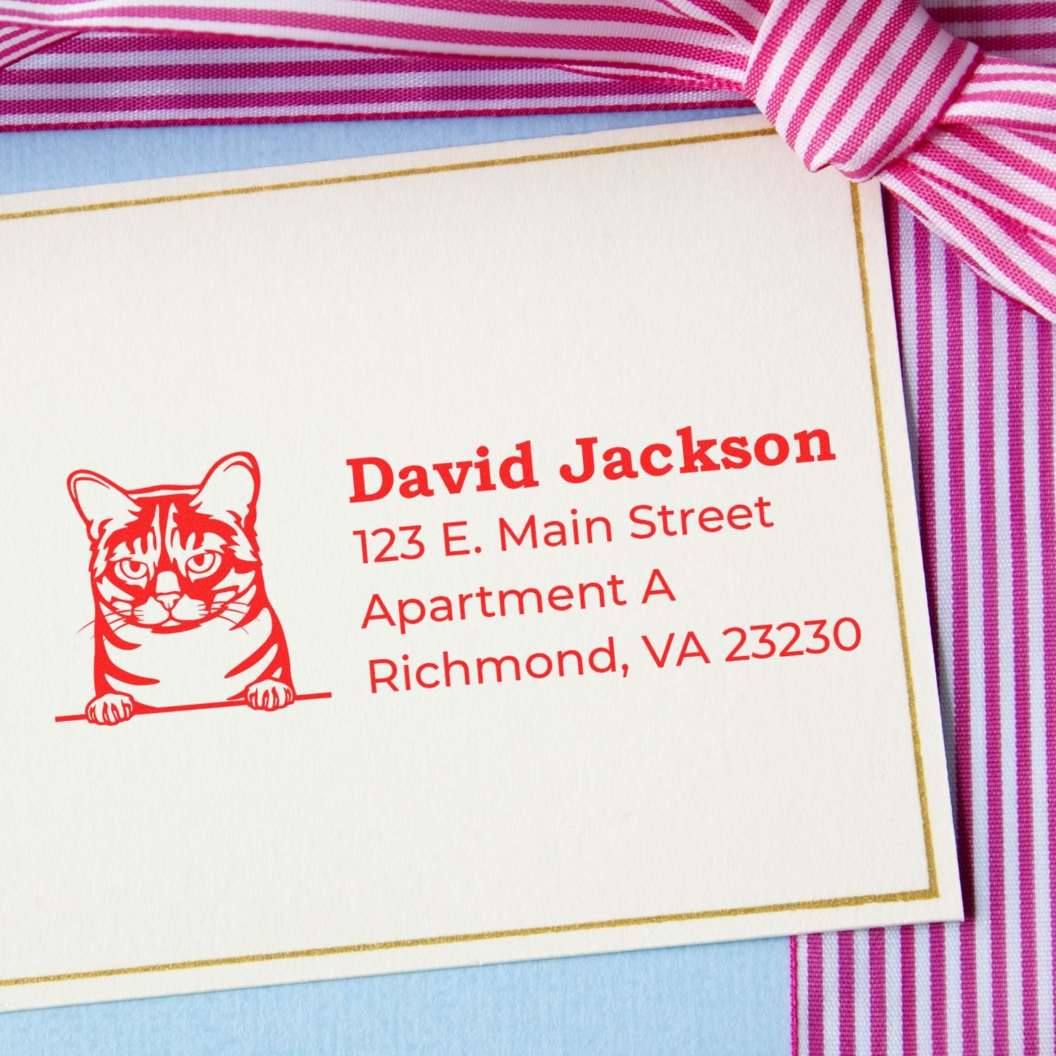 Self-Inking Australian Mist Custom Address Stamp on a card with a red cat design, featuring an address in bold red text. The card is placed on a blue surface with a pink striped ribbon.