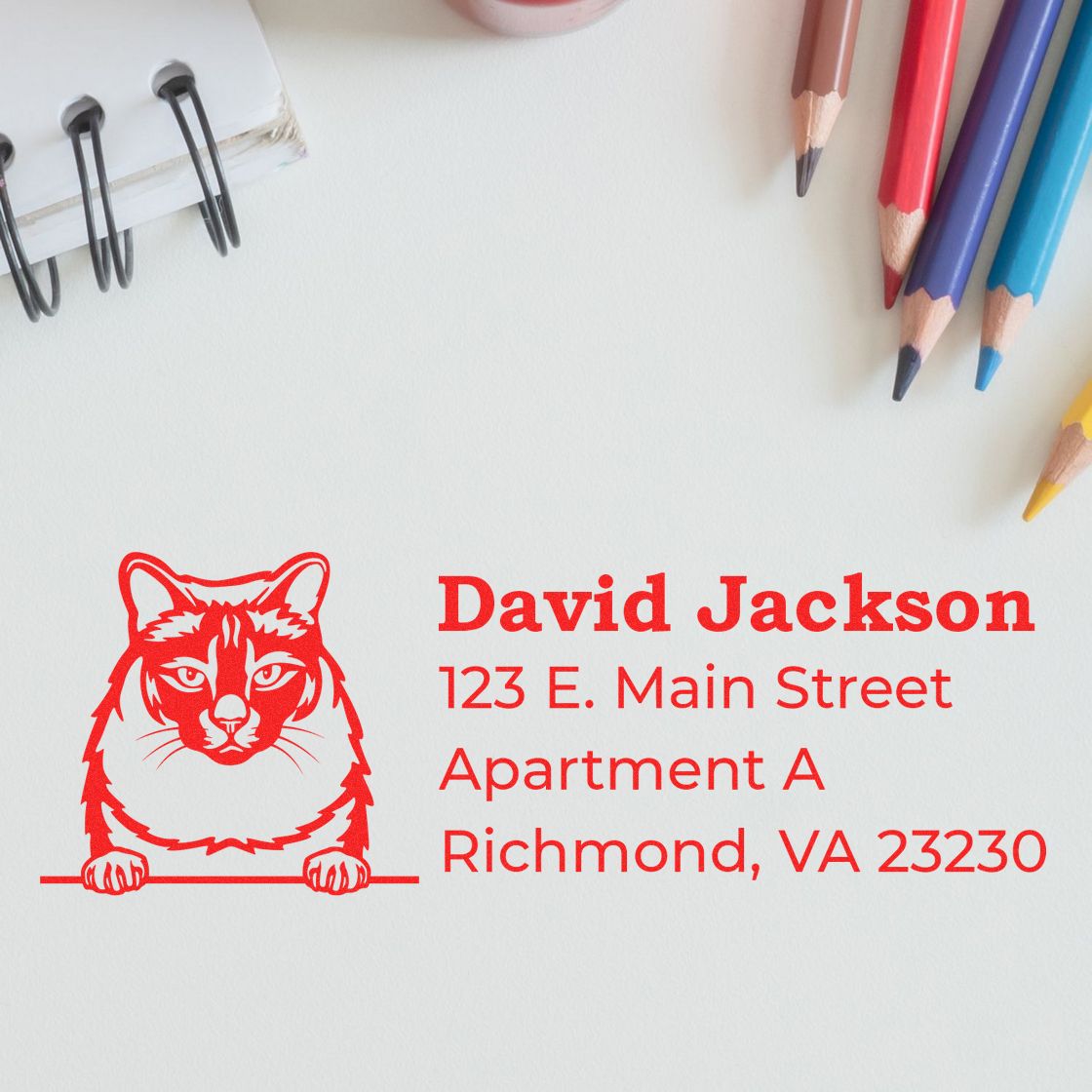 Self-Inking Balinese Custom Address Stamp with red cat design, displaying name and address on white paper, surrounded by colored pencils and a notebook.