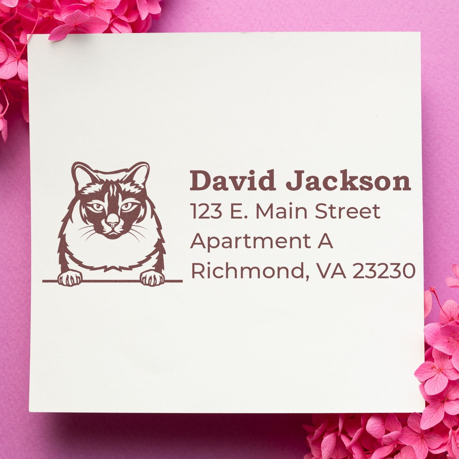 Self-Inking Balinese Custom Address Stamp on white paper with a cat illustration, featuring the name and address in brown ink. Pink background with flower accents.