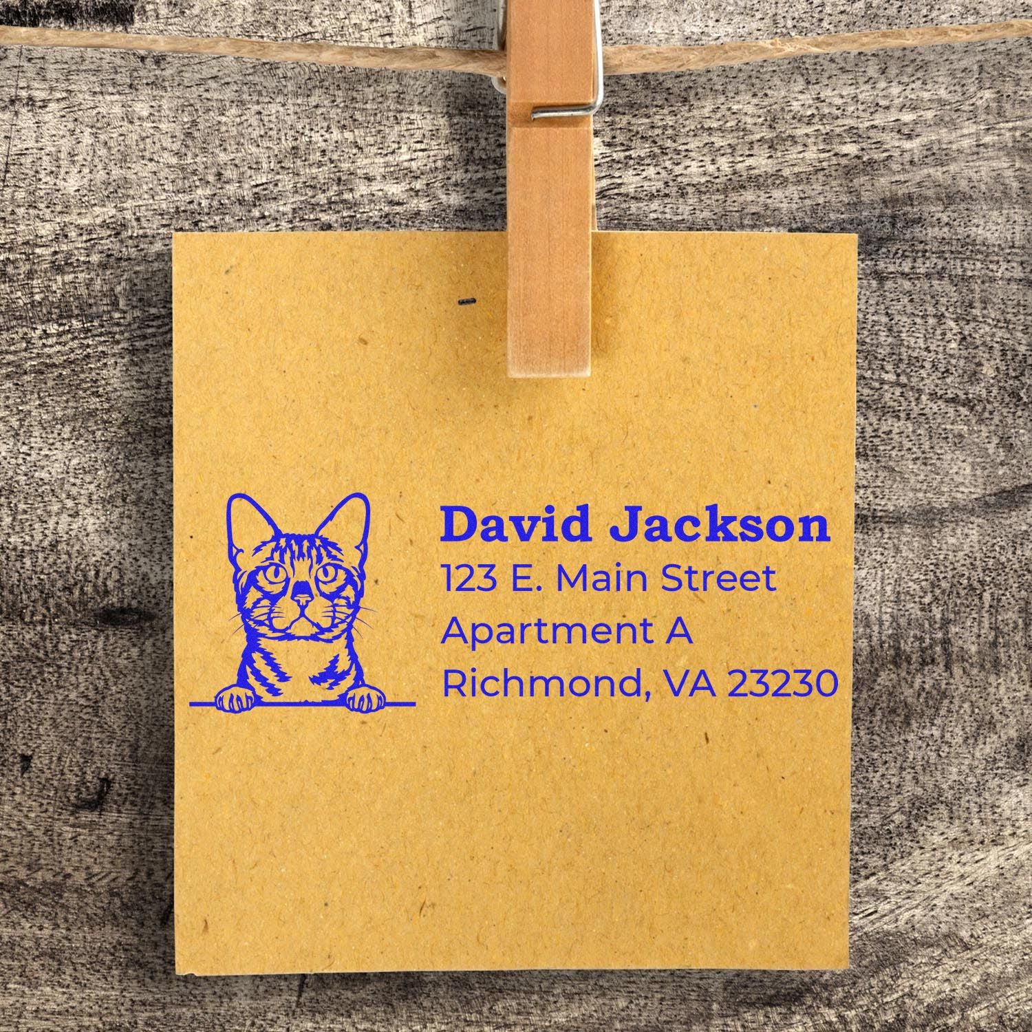 Self-Inking Bengal Custom Address Stamp on brown paper with a cat illustration, blue text showing an address. The paper is clipped to a string against a wooden background.