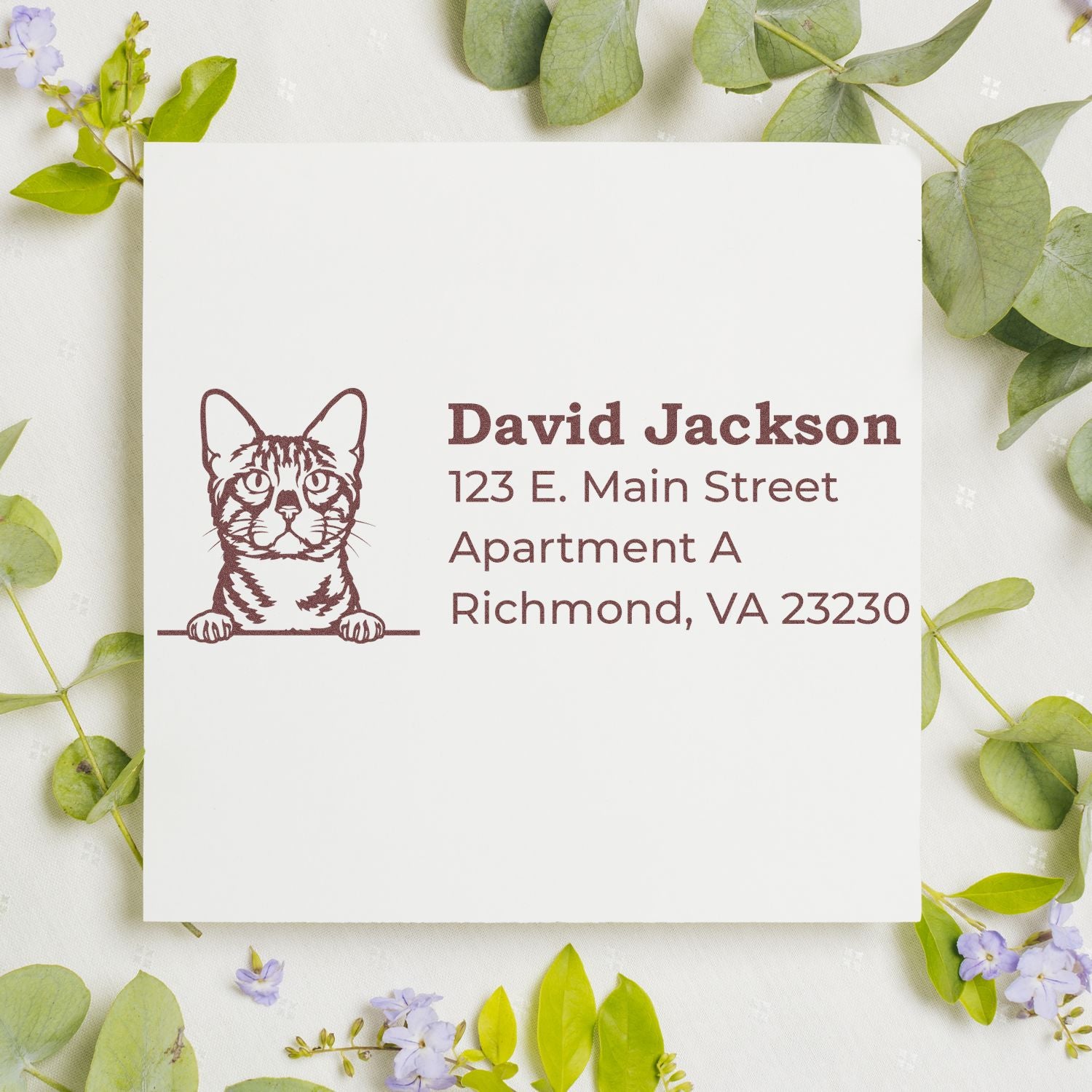 Self-Inking Bengal Custom Address Stamp on white paper with a cat illustration, surrounded by green leaves and small flowers. Text reads: David Jackson, 123 E. Main Street, Apartment A, Richmond, VA 23230.