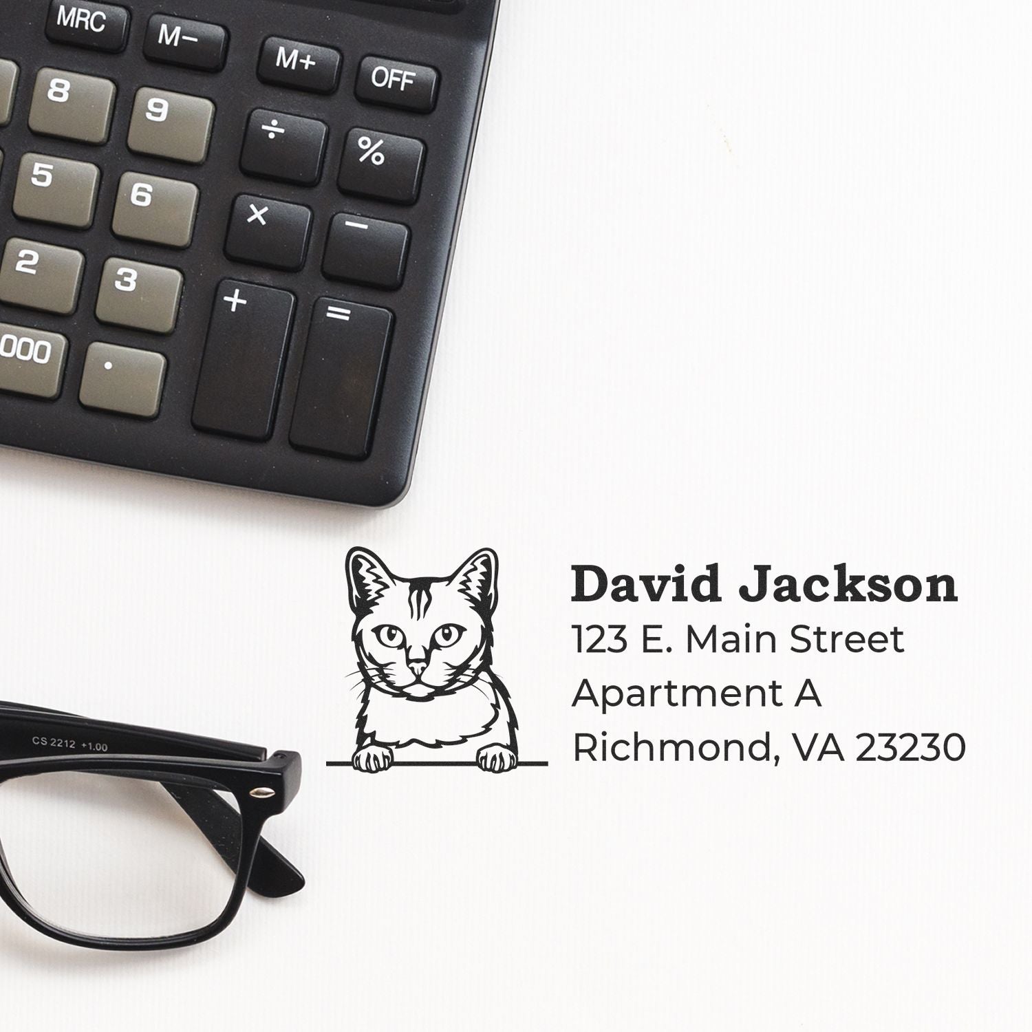 Wood Handle Burmilla Cat Address Stamp on paper with a cat illustration, next to a calculator and glasses. Text reads David Jackson, 123 E. Main Street, Apartment A, Richmond, VA 23230.