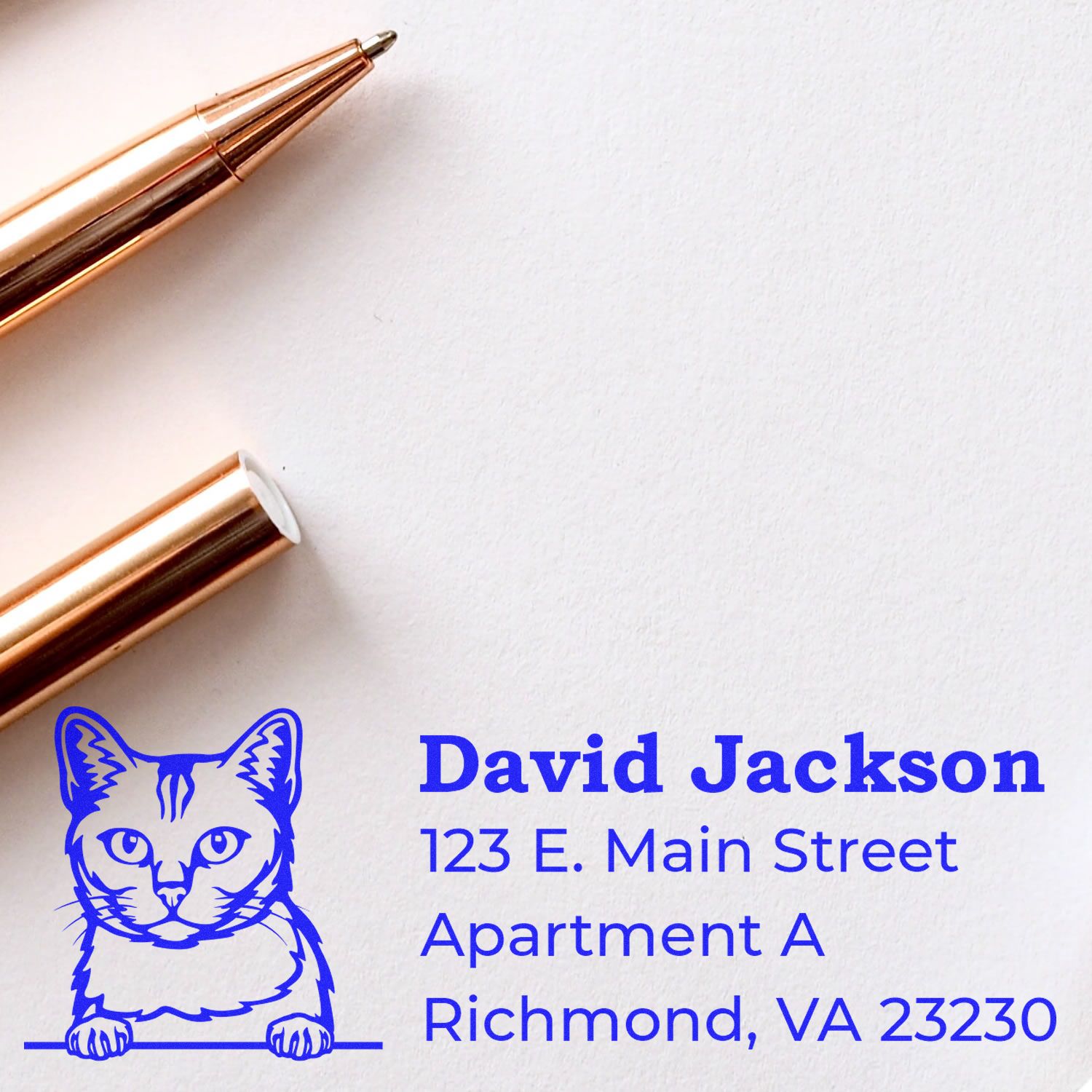 A Self-Inking Burmilla Custom Address Stamp on white paper, featuring a blue cat design and sample address. A gold pen lies nearby, highlighting the stamp's elegant and practical use.