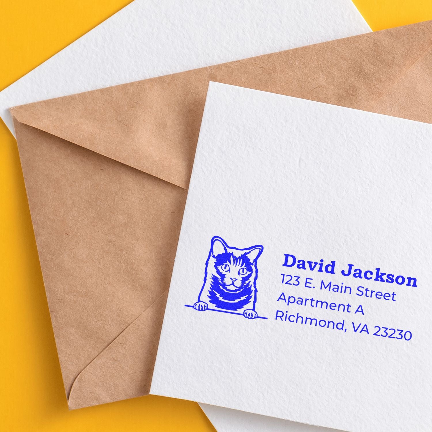 Self-Inking Chartreux Custom Address Stamp on white envelope with blue ink, featuring a cat illustration and address details. Brown envelope and yellow background enhance the presentation.