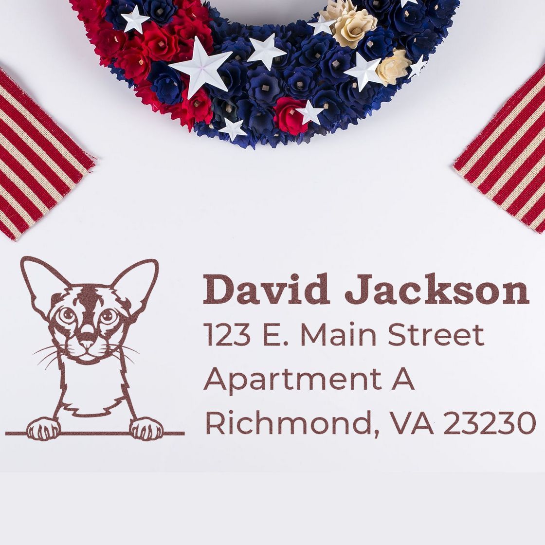 Slim Pre-Inked Colorpoint Shorthair Peeking Cat Return Address Stamp on an envelope with a patriotic wreath and striped ribbons. The stamp features a cat illustration and address details.