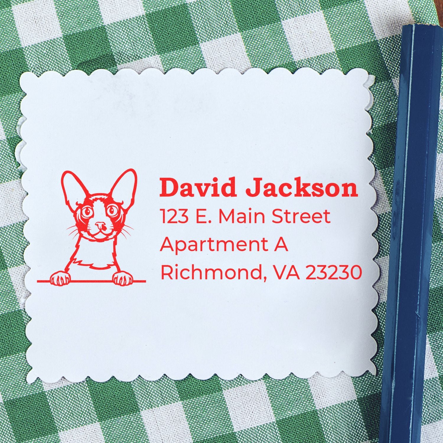 PSI Pre-Inked Peeking Cornish Rex Cat Personalized Address Stamp on scalloped paper, featuring a red cat illustration and sample address, placed on a green checkered tablecloth.