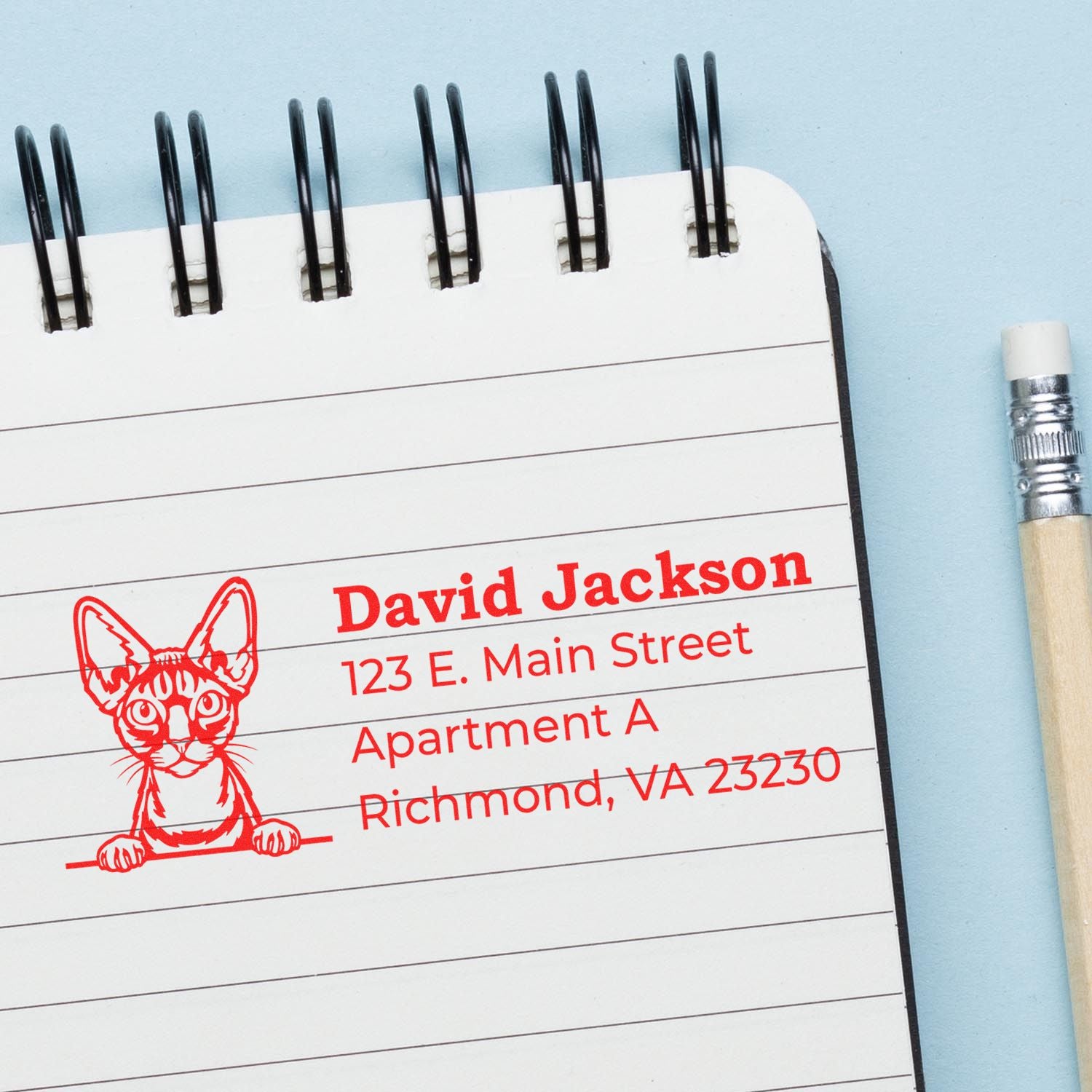 Self-Inking Devon Rex Custom Address Stamp on a notepad, featuring a red cat design with sample address details. A pencil lies beside the notepad on a light blue background.