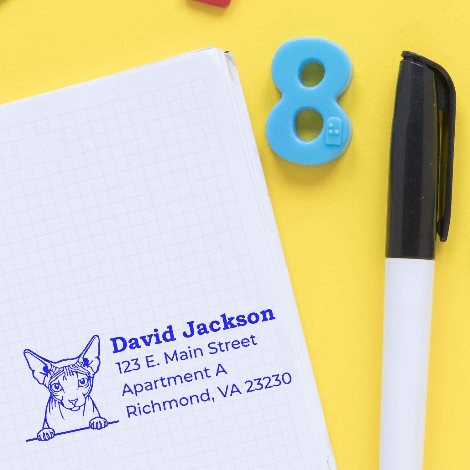 Wood Handle Don Sphynx Cat Address Stamp on a notepad with a blue cat illustration and address details. Nearby are a black pen and a blue number eight on a yellow background.