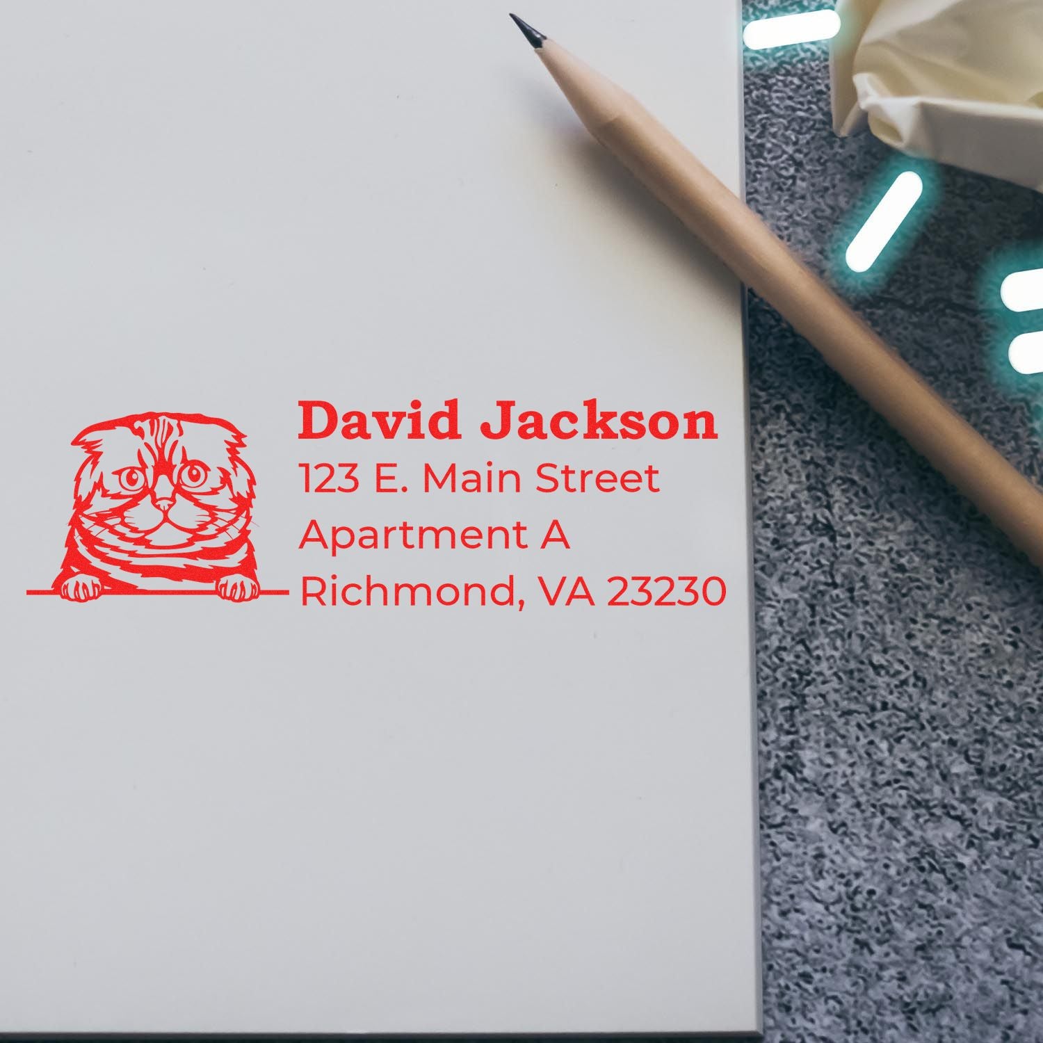 A Self-Inking Foldex Custom Address Stamp on paper, featuring a red cat design and address details, next to a pencil on a textured surface.