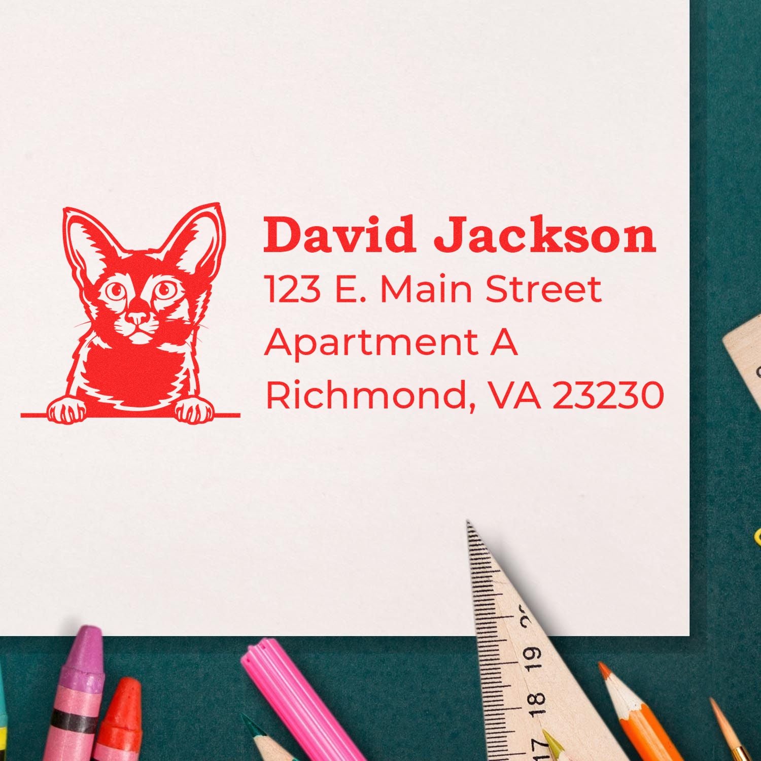 Slim Pre-Inked Javanese Peeking Cat Return Address Stamp on white paper with red ink, featuring a cat illustration and sample address. Surrounded by colorful pencils and a ruler on a green surface.