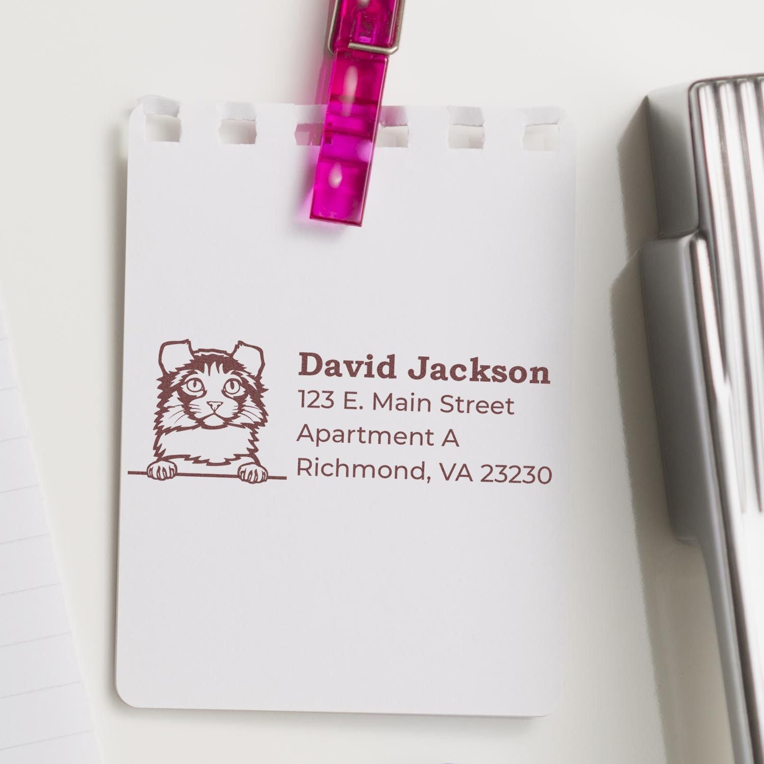 Wood Handle Kinkalow Cat Address Stamp on white paper, featuring a cute cat illustration and sample address. A pink clip holds the paper, with a stapler nearby.