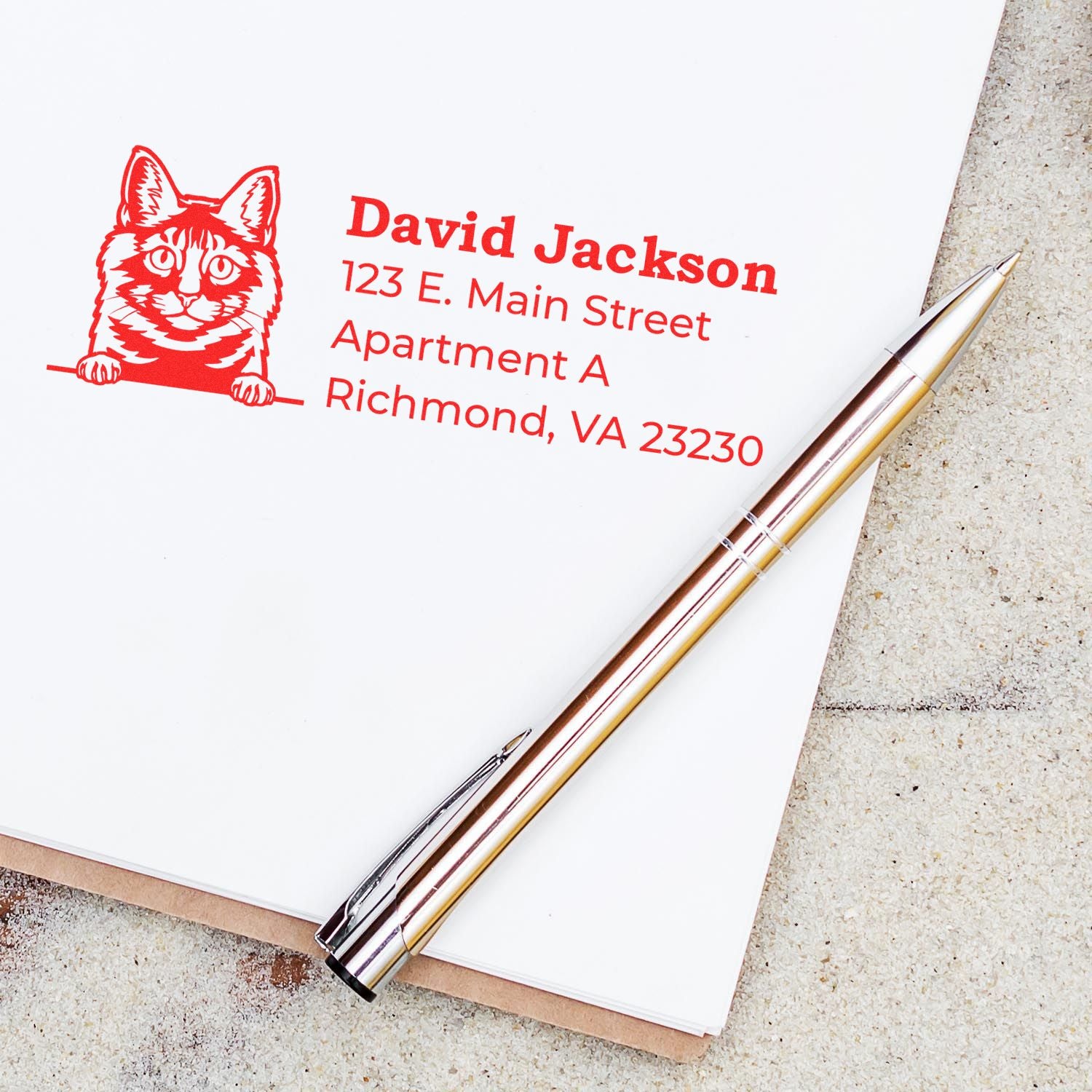 Self-Inking Kurilian Bobtail Custom Address Stamp on paper with a red cat design, showing an address. A pen lies nearby on a textured surface.