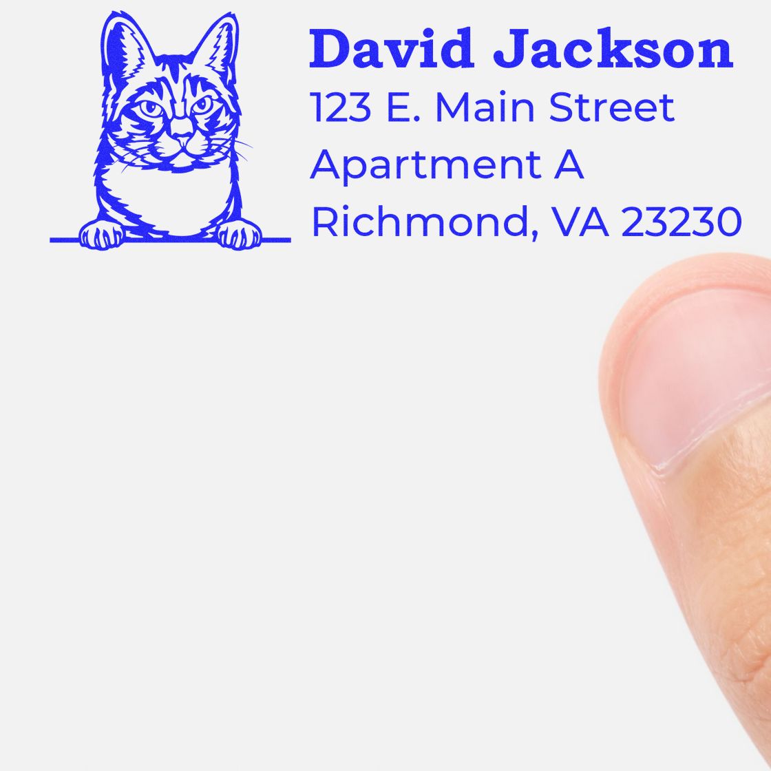 A thumb presses the Self-Inking Manx Custom Address Stamp onto paper, leaving a blue imprint with a cat illustration and sample address: David Jackson, 123 E. Main Street, Apartment A, Richmond, VA 23230.