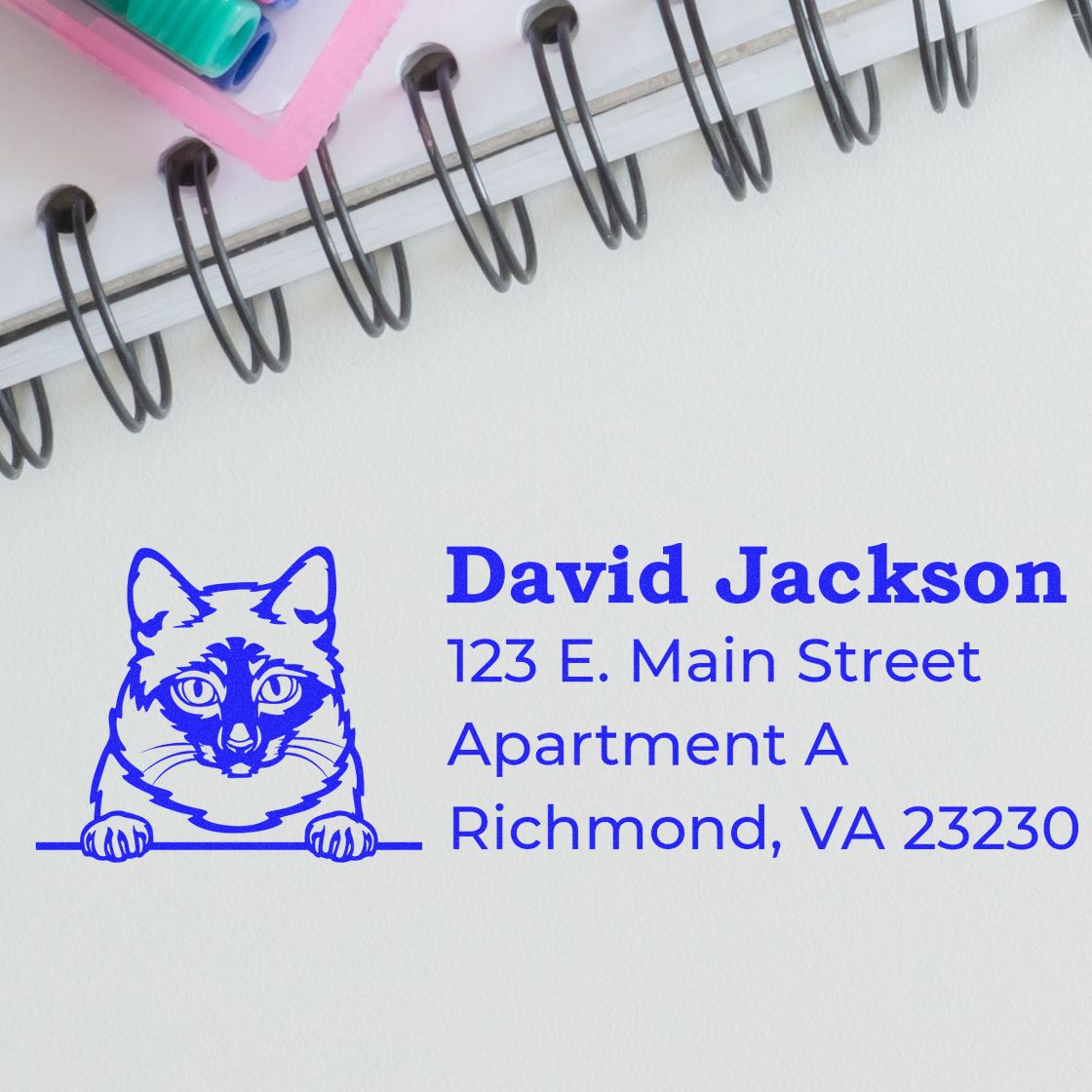 Self-Inking Mekong Bobtail Custom Address Stamp in use on white paper, featuring a blue cat design and sample address. Nearby, a spiral notebook and colorful pens add a touch of office decor.