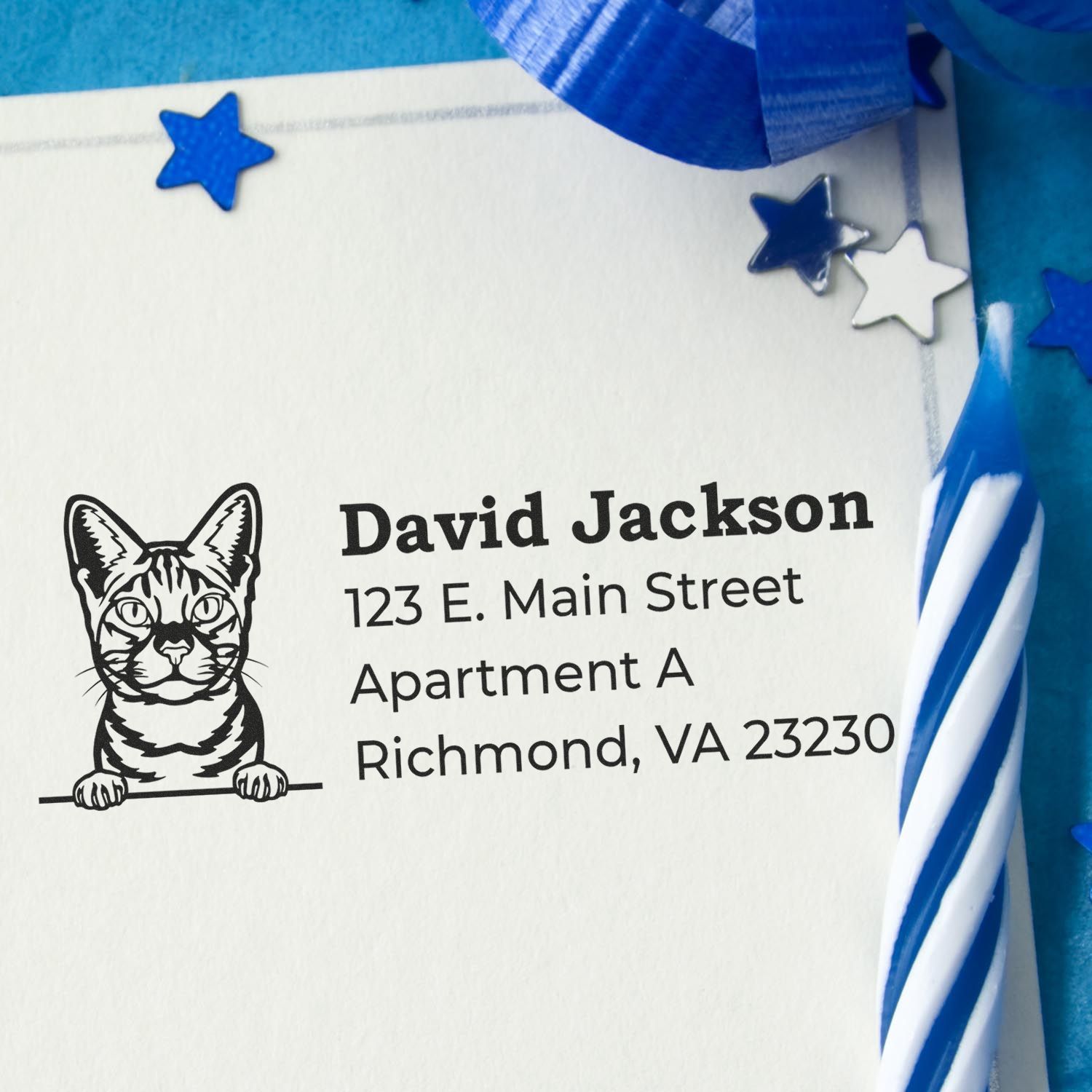 Slim Pre-Inked Ocicat Peeking Cat Return Address Stamp on an envelope with a blue candle and star confetti. The stamp features a cute cat design above the address text.