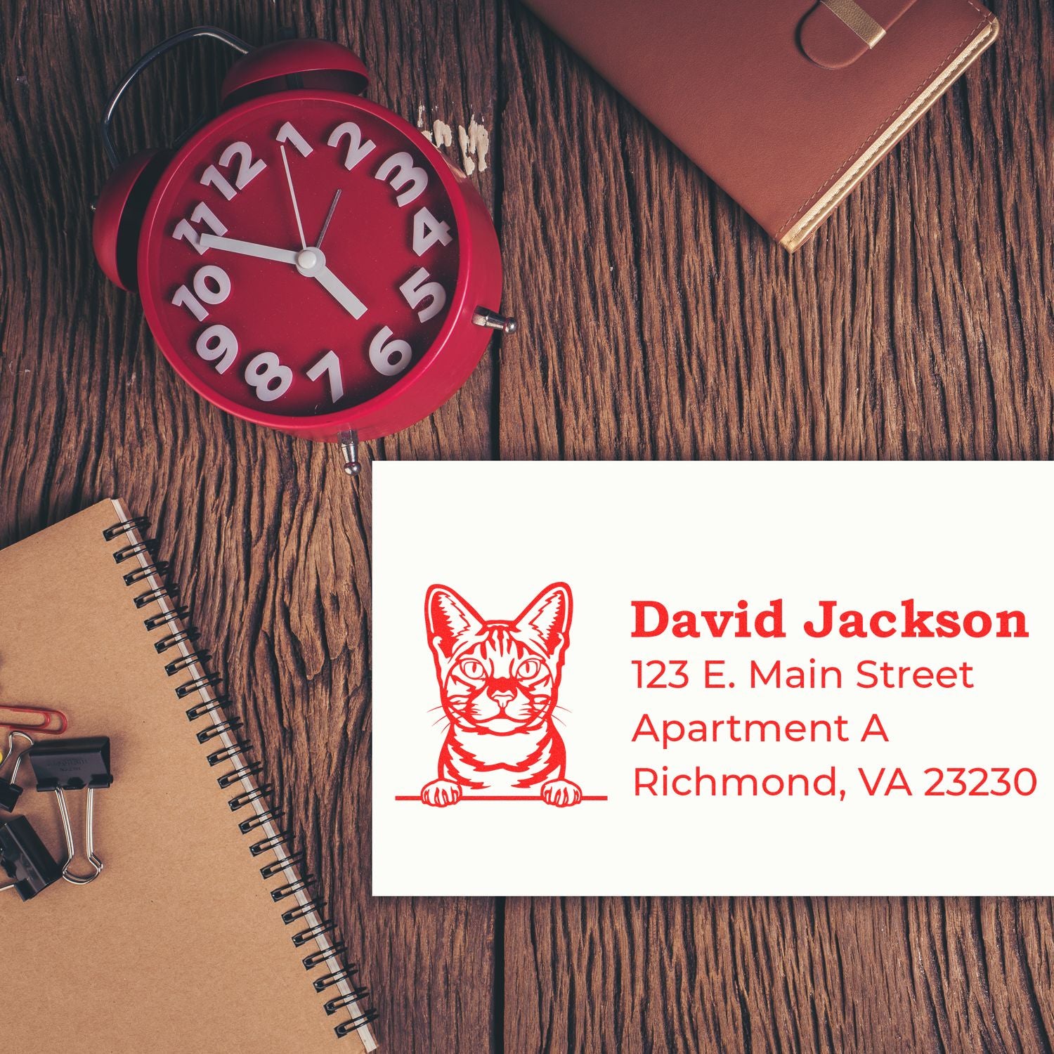 A Self-Inking Ocicat Custom Address Stamp on a white card with a red ocicat design, next to a red clock, notebook, and binder clips on a wooden surface.