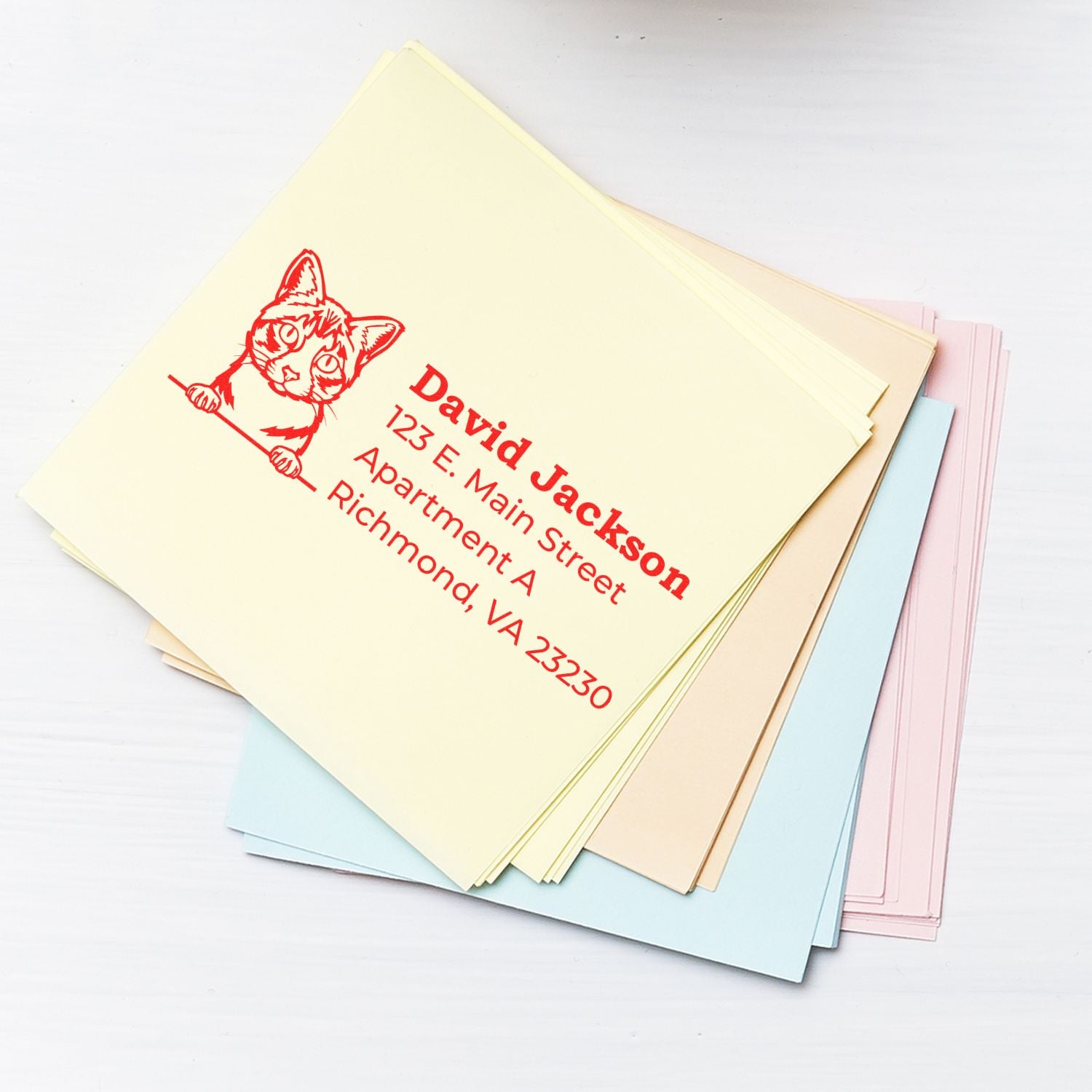 Stack of pastel envelopes stamped with the Self-Inking Ojos Azules Custom Address Stamp, featuring a cat design and red text.