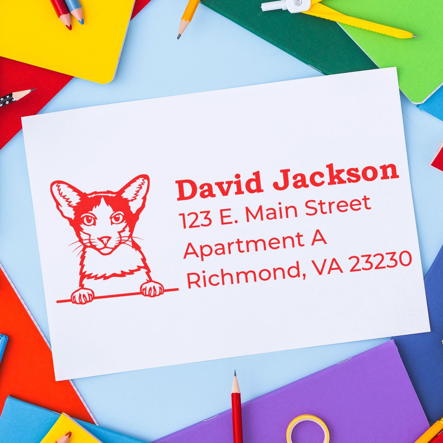 Self-Inking Oriental Bicolor Custom Address Stamp on white paper with a red cat design, surrounded by colorful stationery, displaying an address in bold red text.