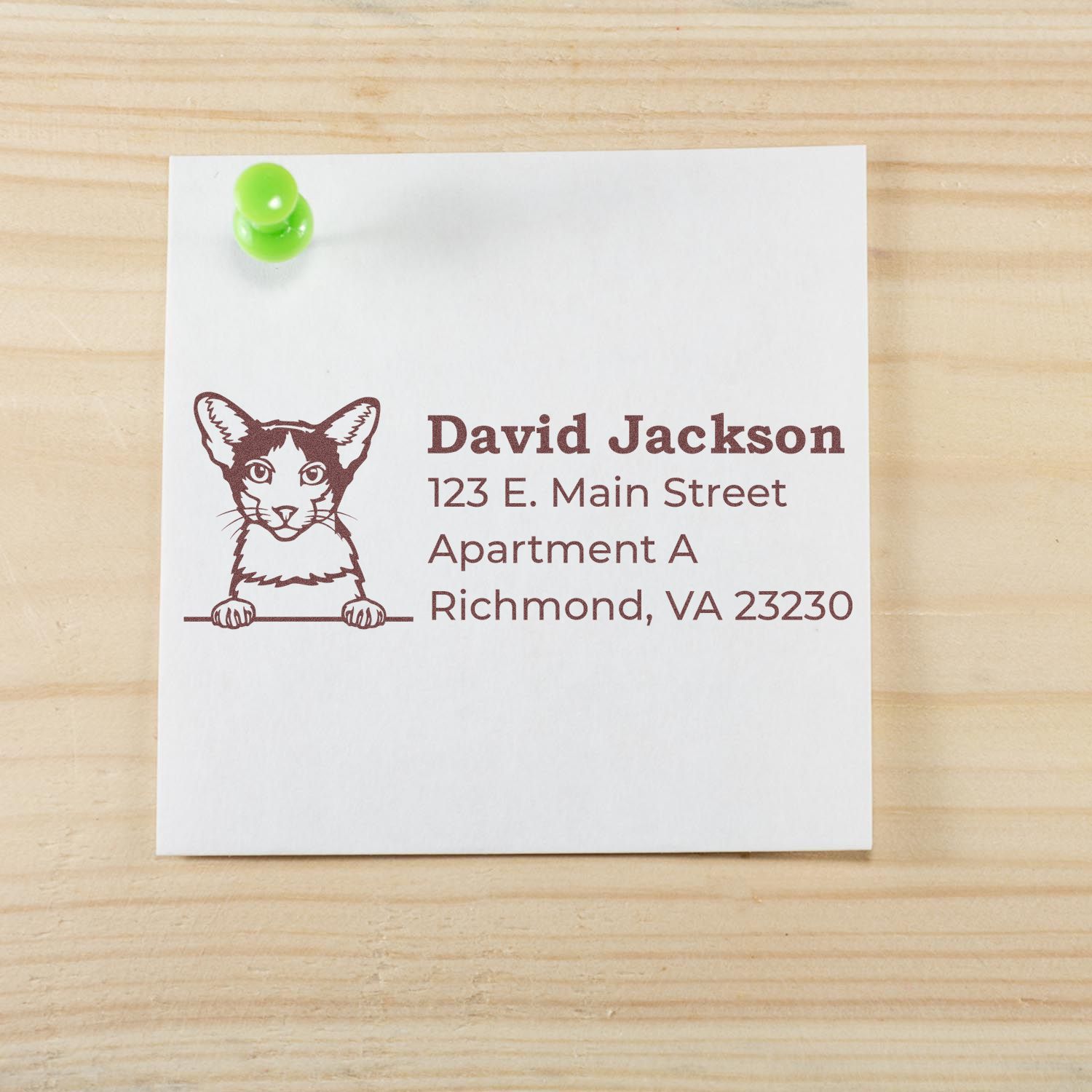 Self-Inking Oriental Bicolor Custom Address Stamp on a note with a cat illustration, displaying an address. The note is pinned to a wooden surface with a green pushpin.