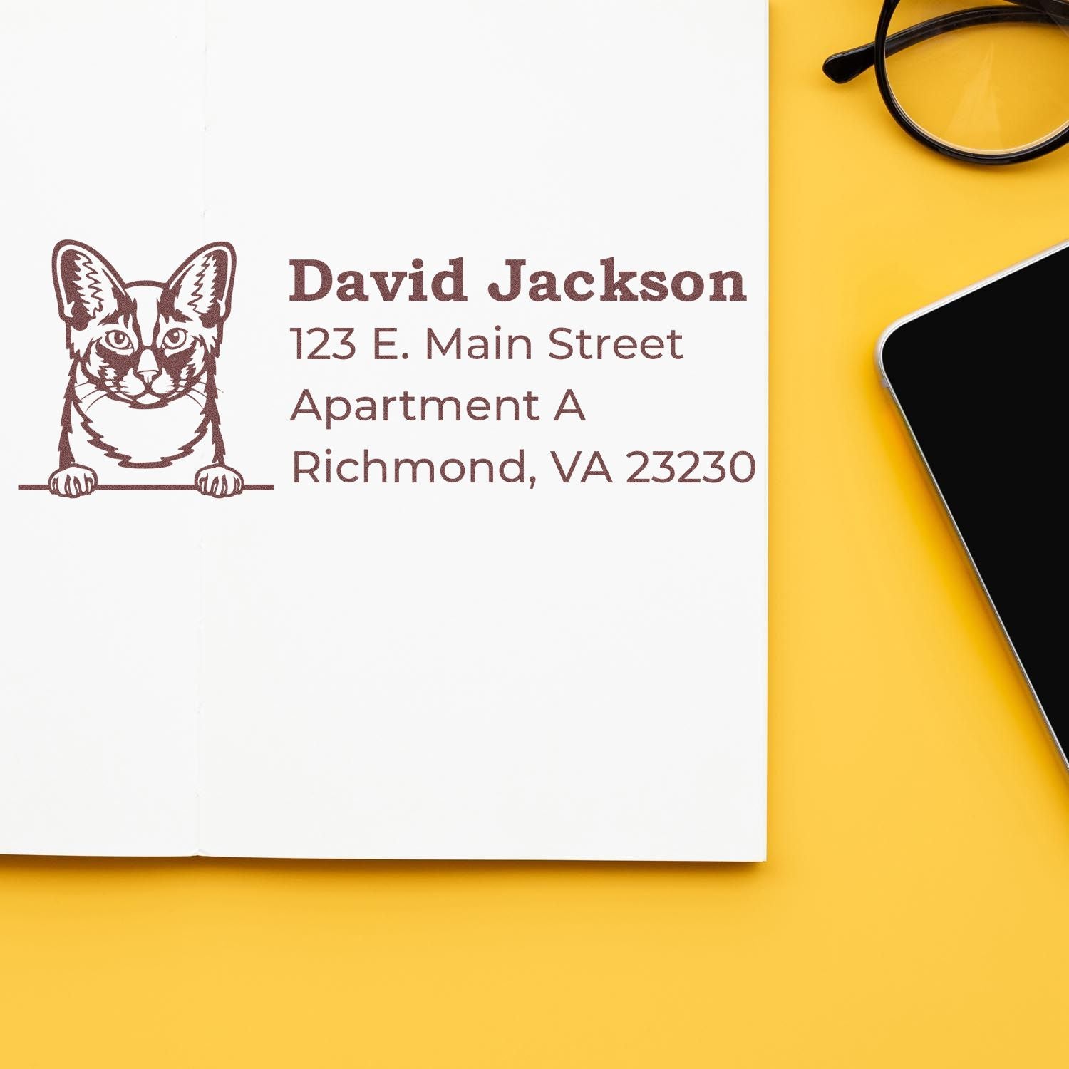 Slim Pre-Inked Oriental Longhair Peeking Cat Return Address Stamp on a white card with a cat illustration, next to glasses and a smartphone on a yellow surface.