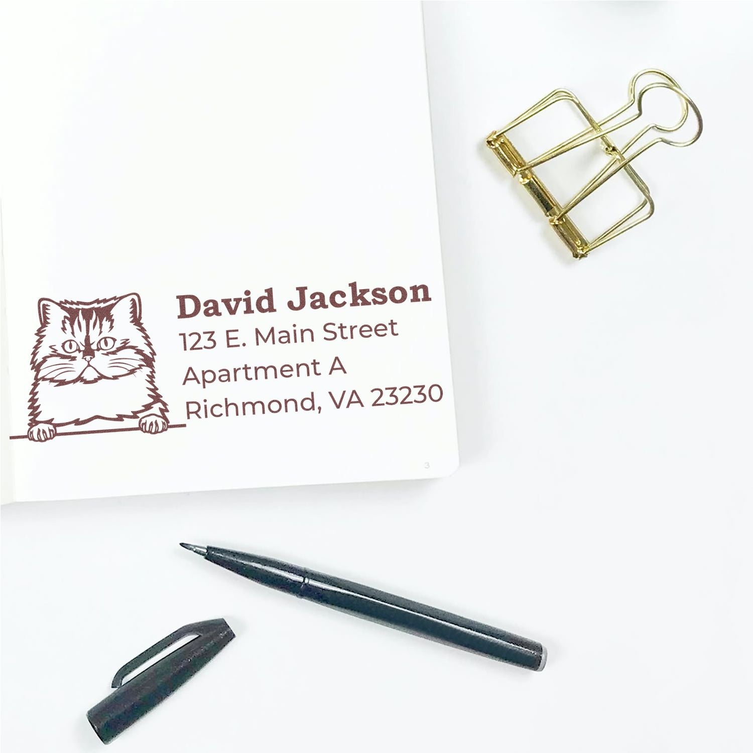 Self-Inking Persian Custom Address Stamp featuring a cat design, shown on a white paper with a pen and gold binder clip nearby.