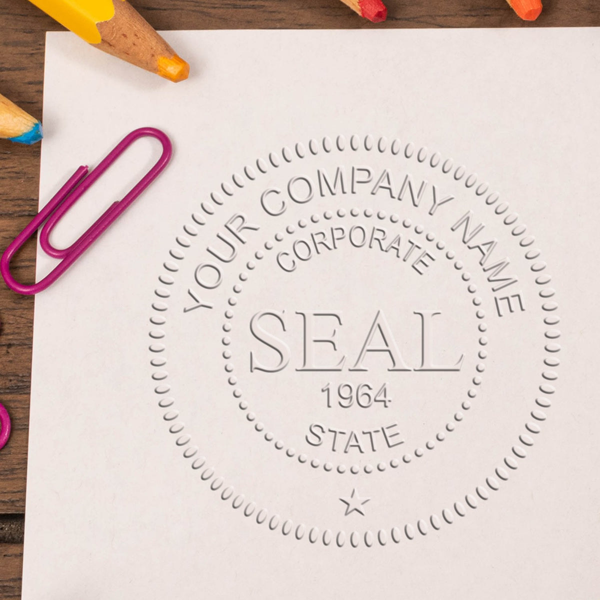 Custom corporate seal stamp embossed in gray on white paper, with a purple paperclip and colored pencils on wood.
