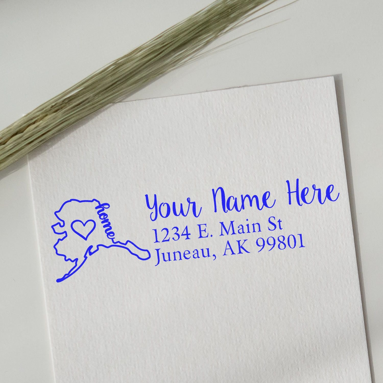 PSI Pre-Inked Personalized Alaska State Love Address Stamp on white paper, featuring a blue Alaska outline with a heart and custom address text, next to a sprig of dried grass.