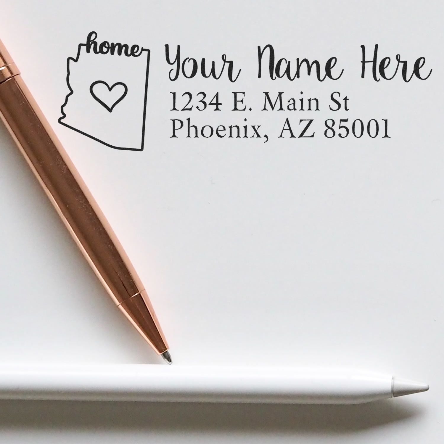 Arizona Self-Inking State Love Address Stamp on white paper with a rose gold pen and a white pen, featuring a heart and 'home' text inside the Arizona state outline, alongside sample address text.